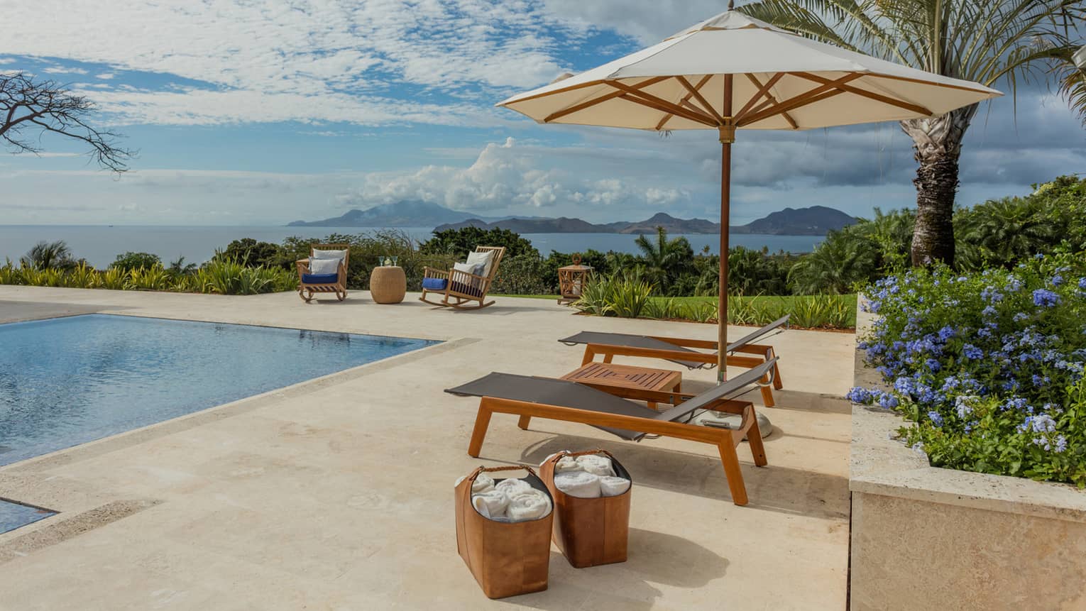 Two lounge chairs under patio umbrella, baskets with towels by outdoor pool