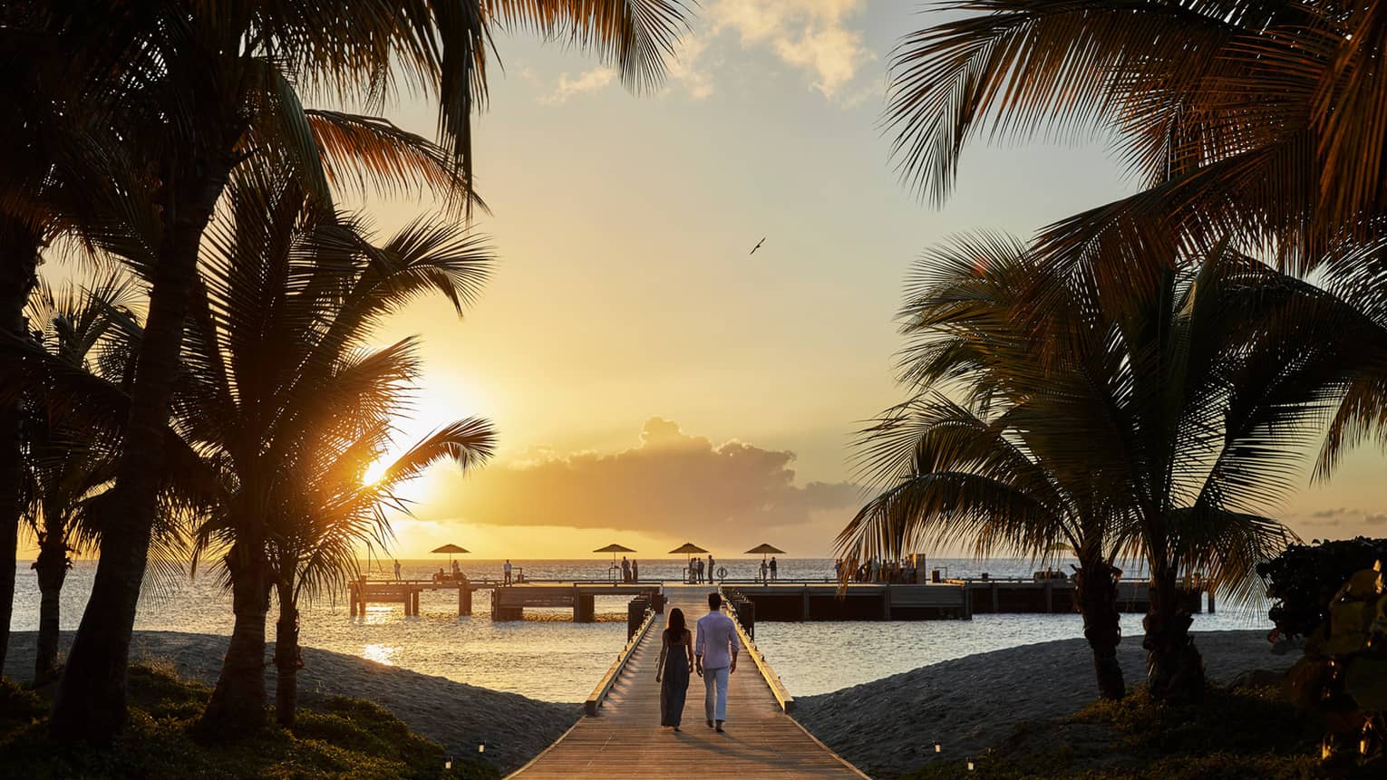 Silhouettes of palm trees, couple walks along boardwalk toward pier at sunset