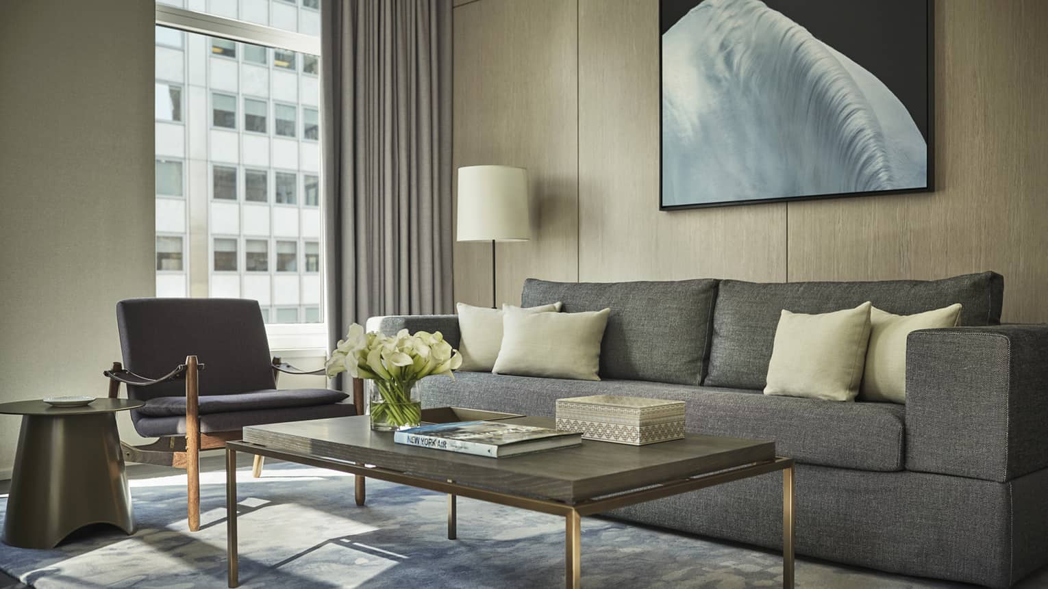 Hudson Suite long grey sofa, table with white flowers, books, armchair by window, building view