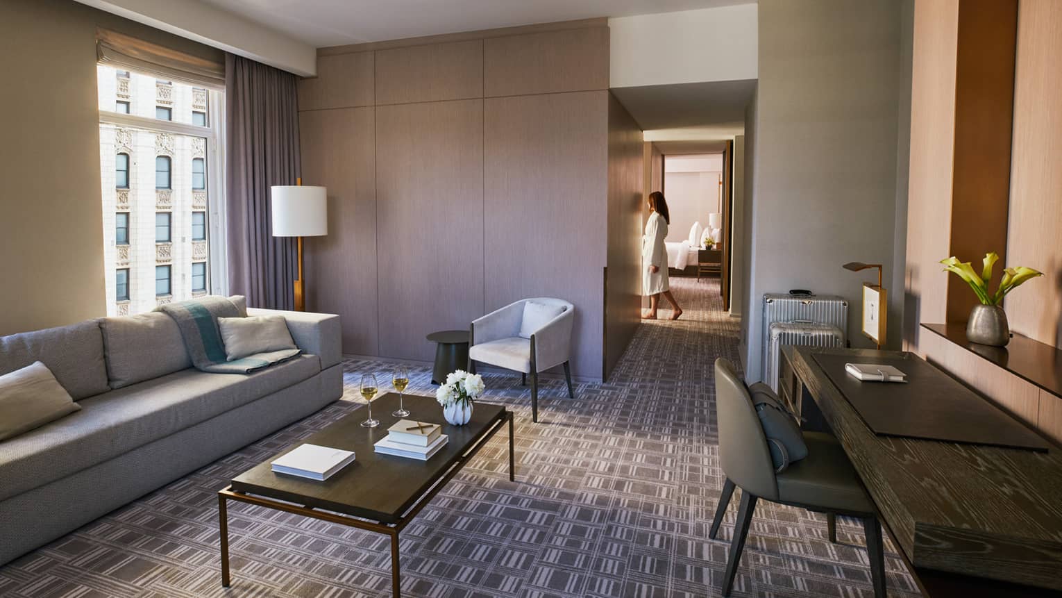 Luxury hotel suite with a gray sofa, coffee table, and desk, featuring a person in a bathrobe walking toward the bedroom area