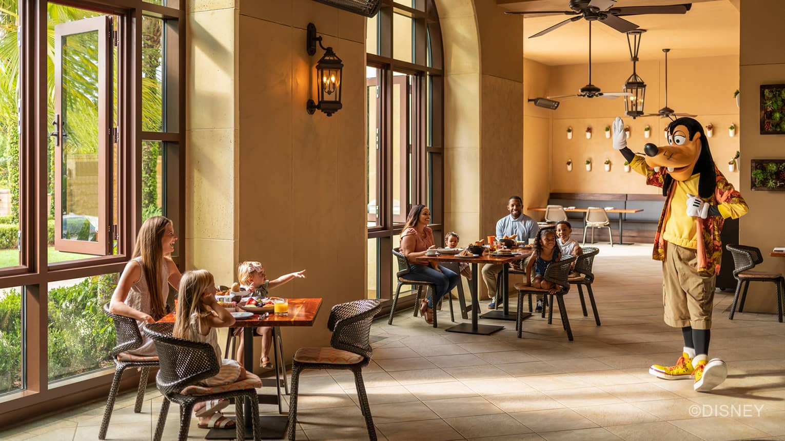 Sunlight filters into a restaurant where two families with young children smile at Goofy as he walks over and waves at them.