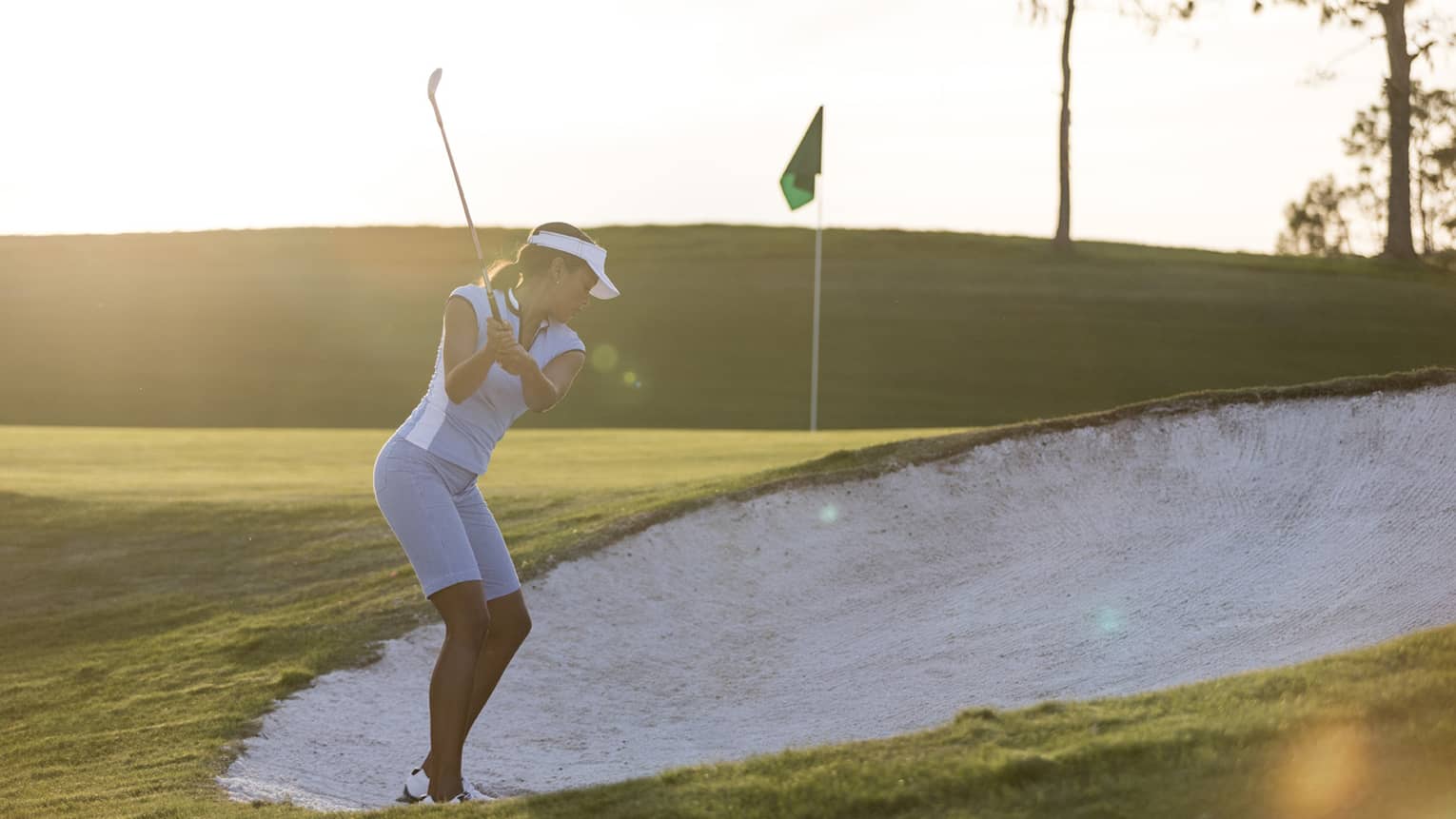 Woman swings golf club in sand trap on green at sunrise