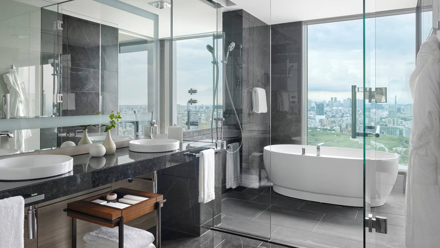 Luxurious bathroom with double-sink vanity area separated by a glass partition from the freestanding bathtub with a panoramic city view