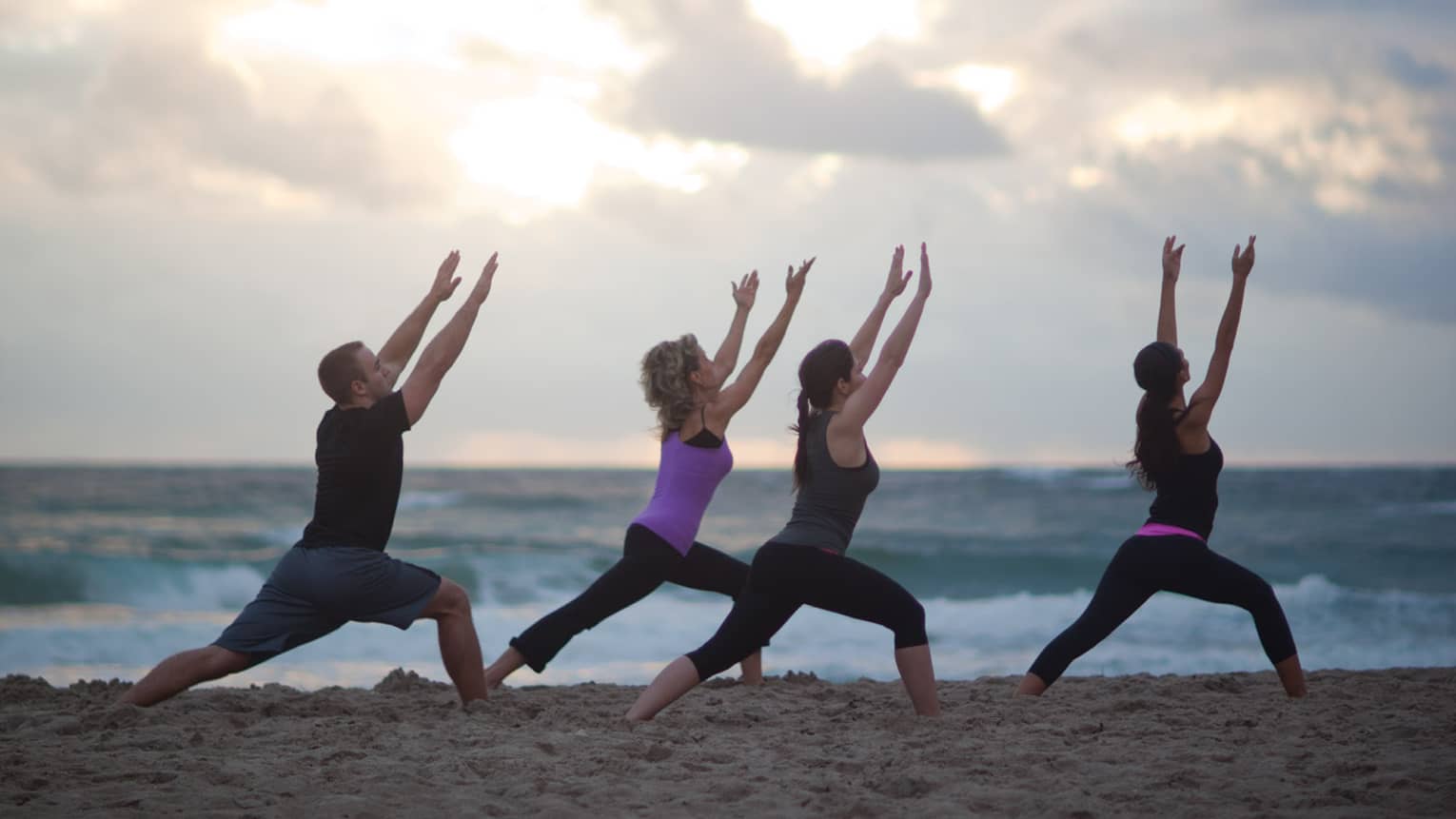 Man and three women in workout clothes do yoga stretches on a beach 