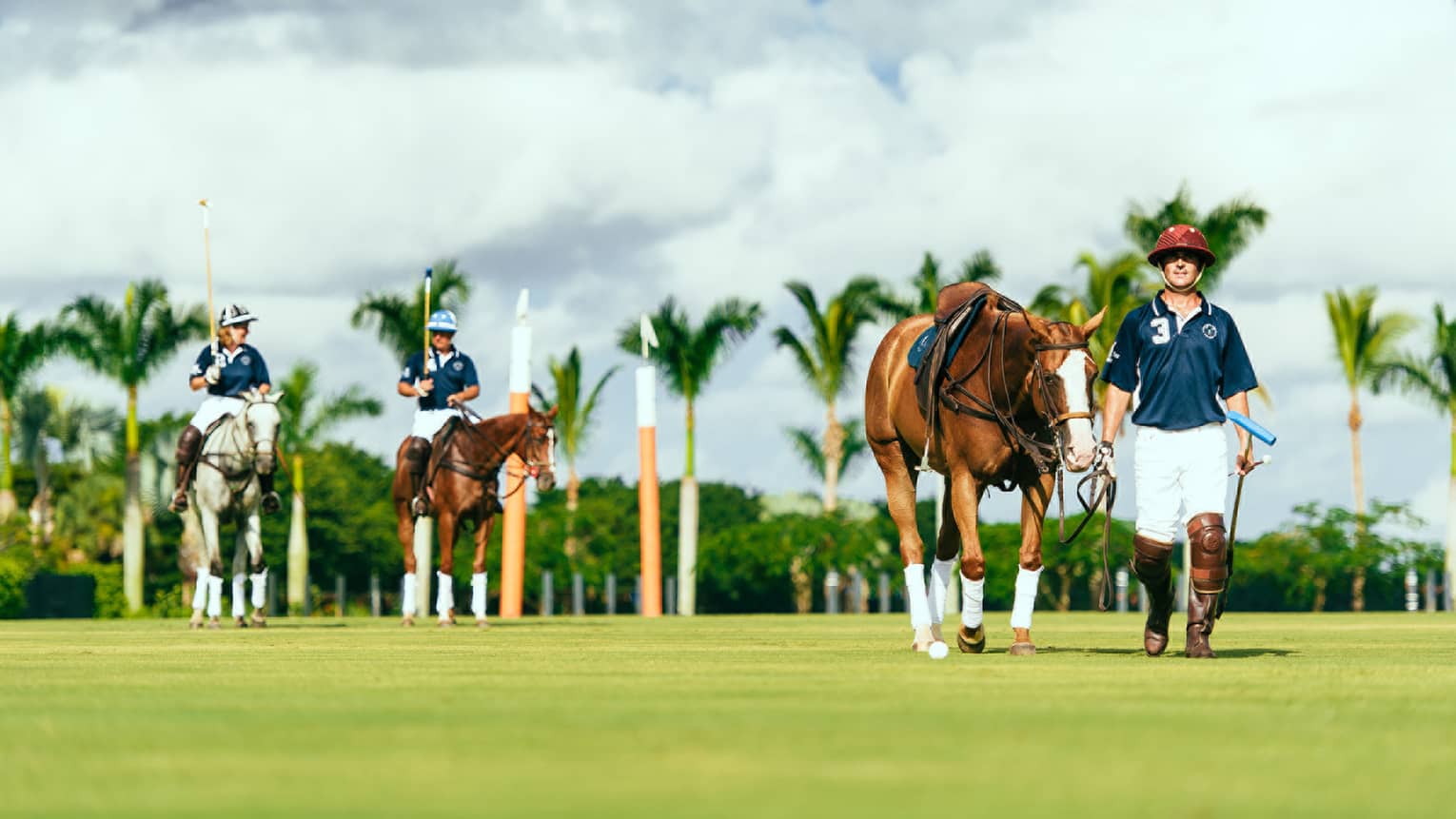A polo player leads his horse forward, while two other players converse while riding their horses behind the walking player. 