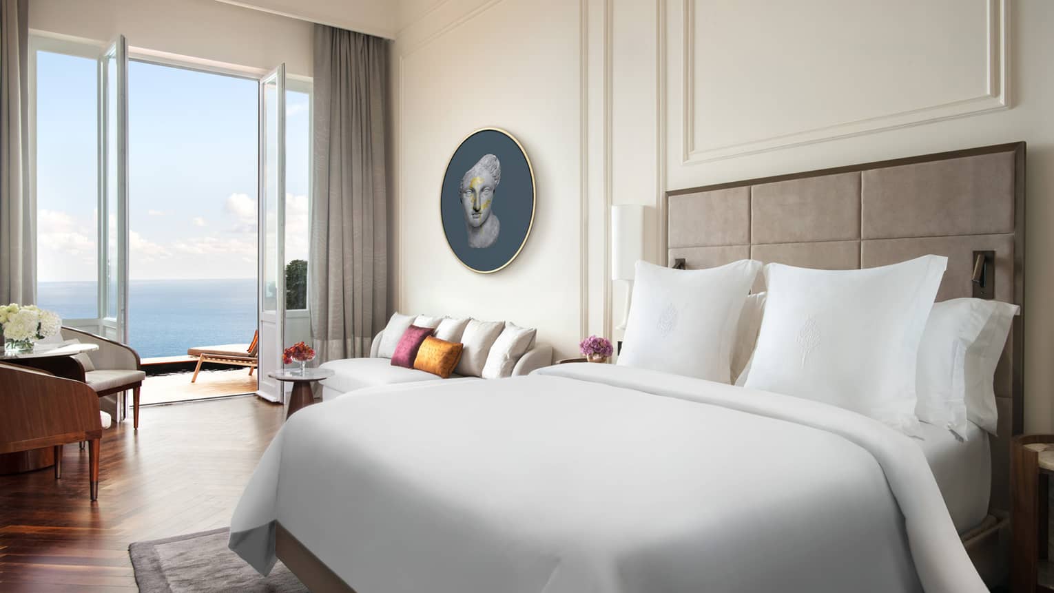 Hotel room with white king bed, white walls with crown molding, wood floors, walk-out sea-view balcony