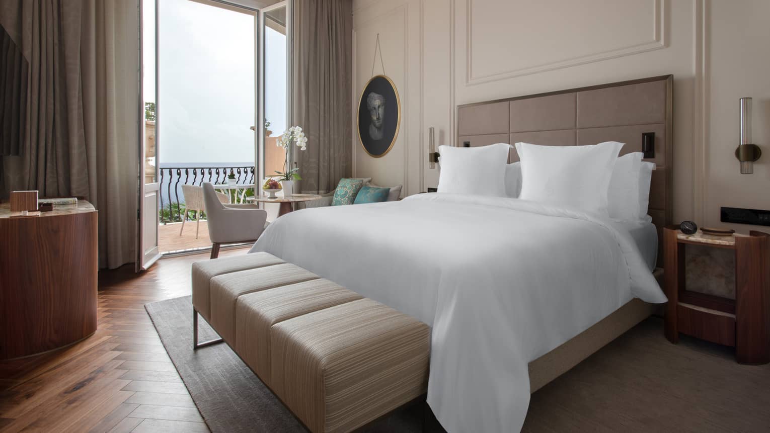 Hotel room with white king bed, tan leather bench, small balcony