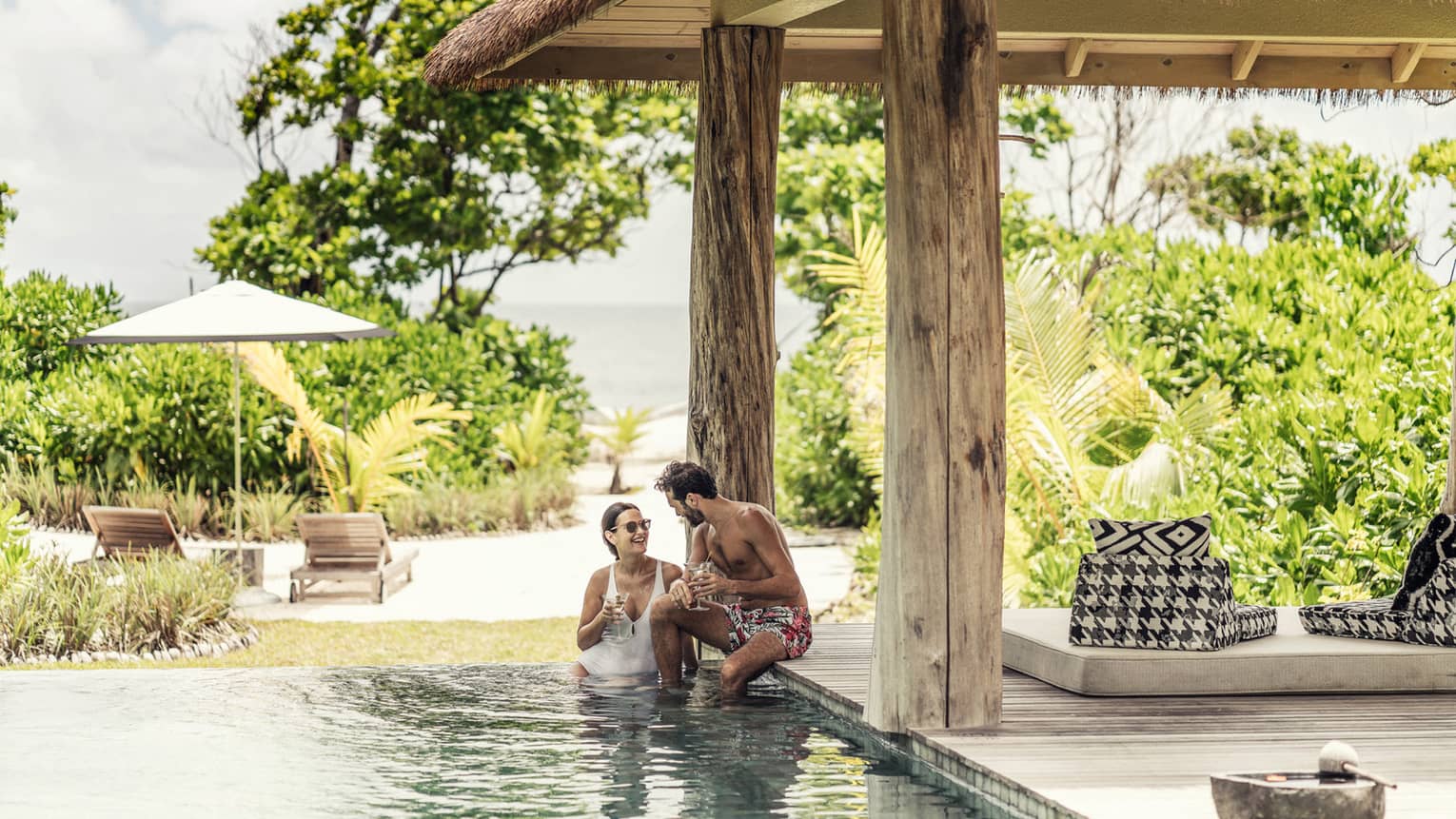 Couple in villa pool under thatched roof and view of tropical foliage