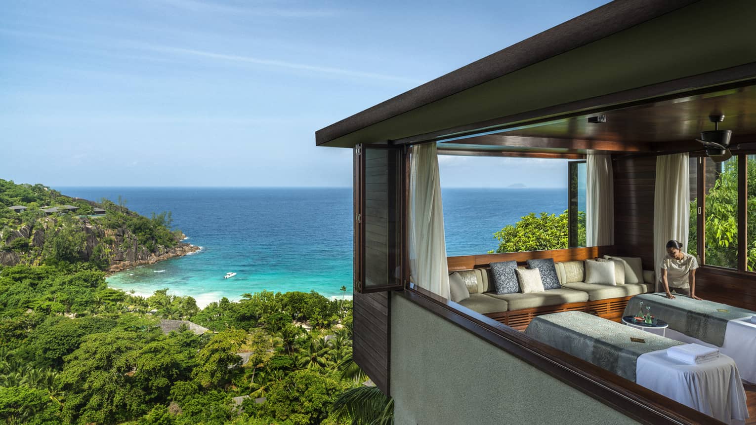 Woman smoothes sheet on side-by-side massage tables in spa room high above trees, ocean