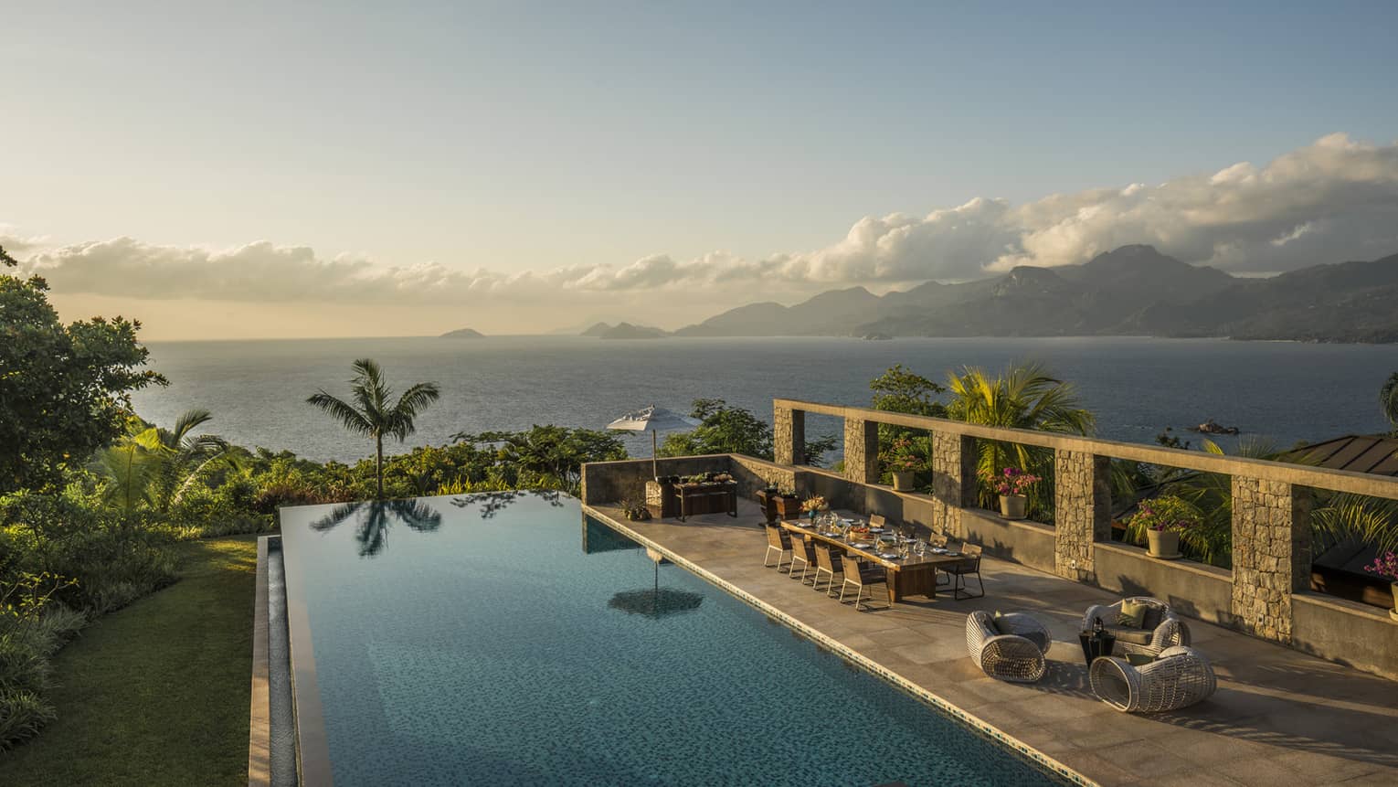 Infinity pool overlooking ocean with palm trees, table, chairs and lounge area 
