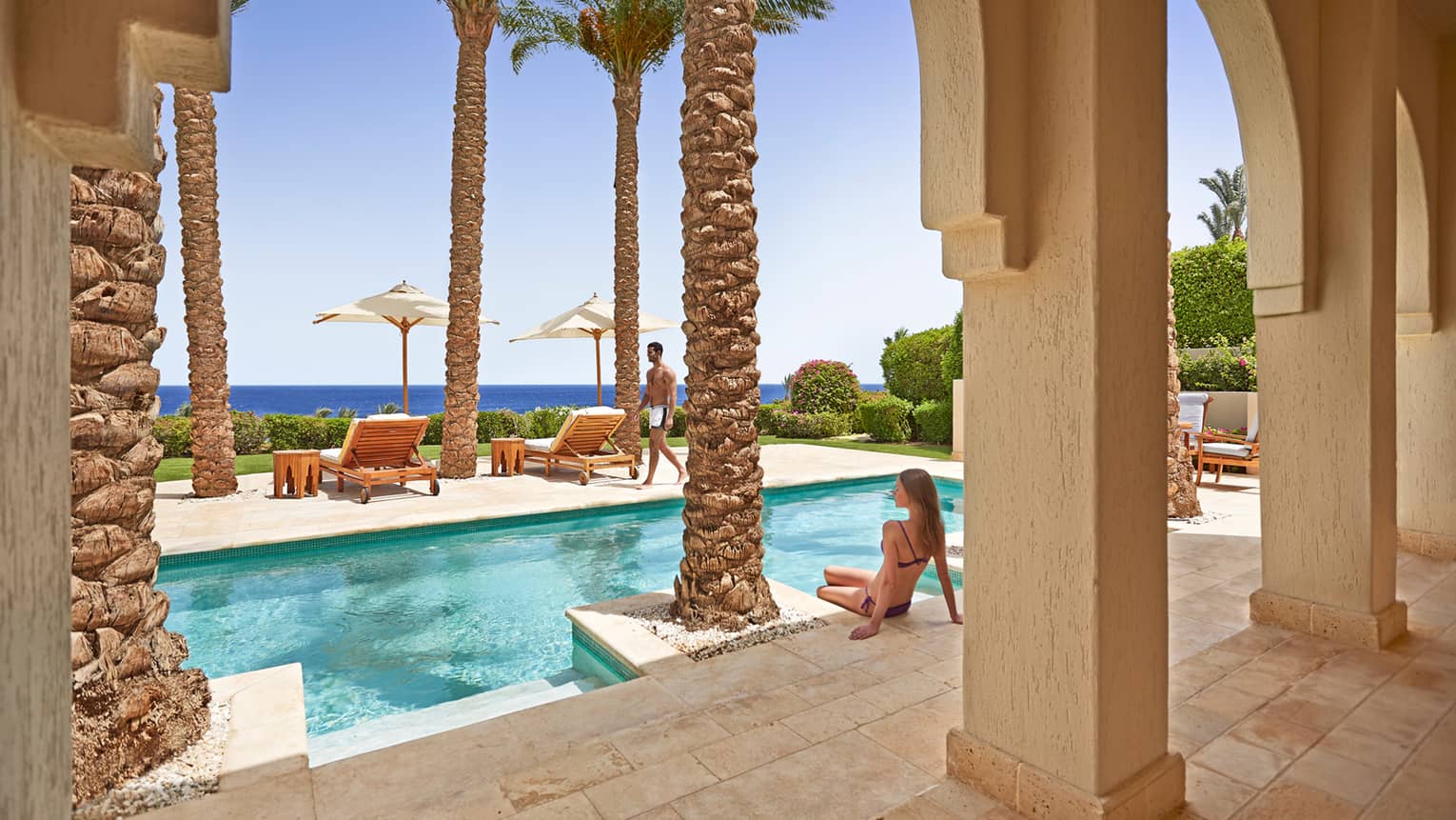 Woman in bikini sits on edge of outdoor swimming pool under palm trees, clay columns