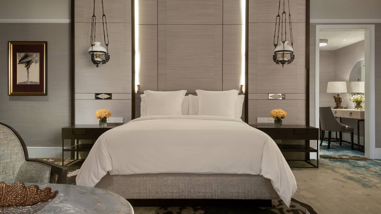 A stylish bedroom with a large bed adorned in white linens, flanked by modern hanging lamps. Nightstands hold vases of yellow roses, and a vanity is visible in the adjacent room.
