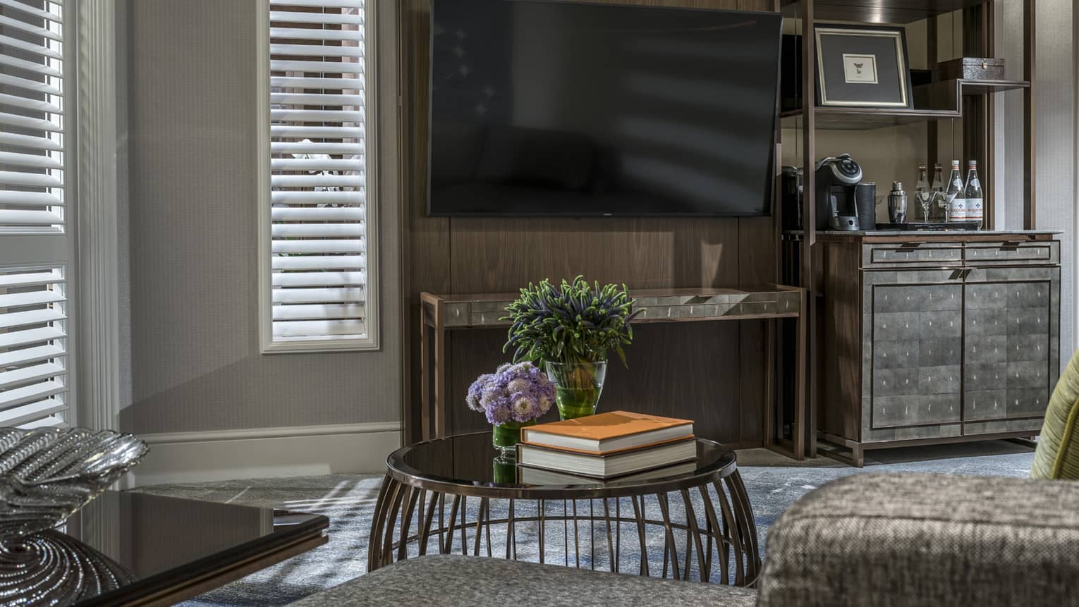 A living area featuring a large wall-mounted TV,  sleek wooden cabinet with a coffee machine, and a round table holding flowers and books. Shuttered windows allow soft light to filter in.
