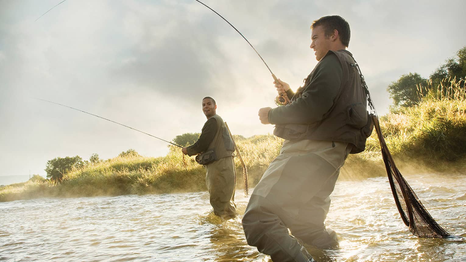 Two people fly fishing in a river, standing knee-deep in the water, surrounded by mist and greenery.