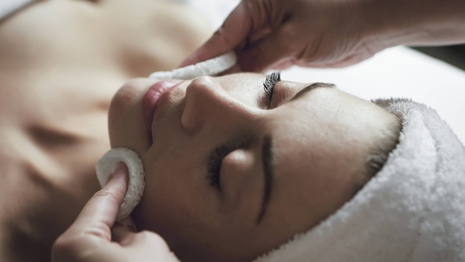 Woman with eyes closed, hair wrapped in towel, as spa attendant rubs cheeks with white pads