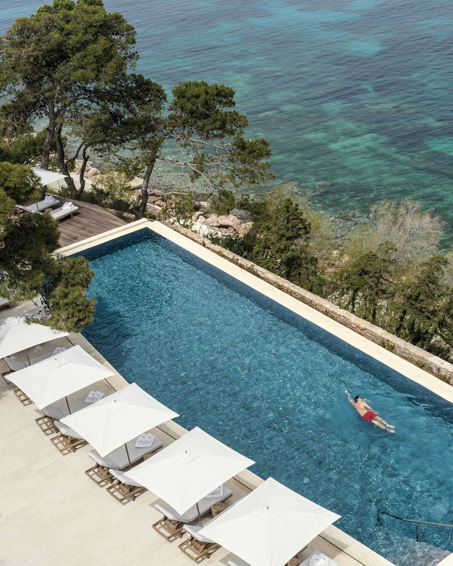 Man in red swim shorts swimming in infinity pool overlooking ocean and white cabanas 