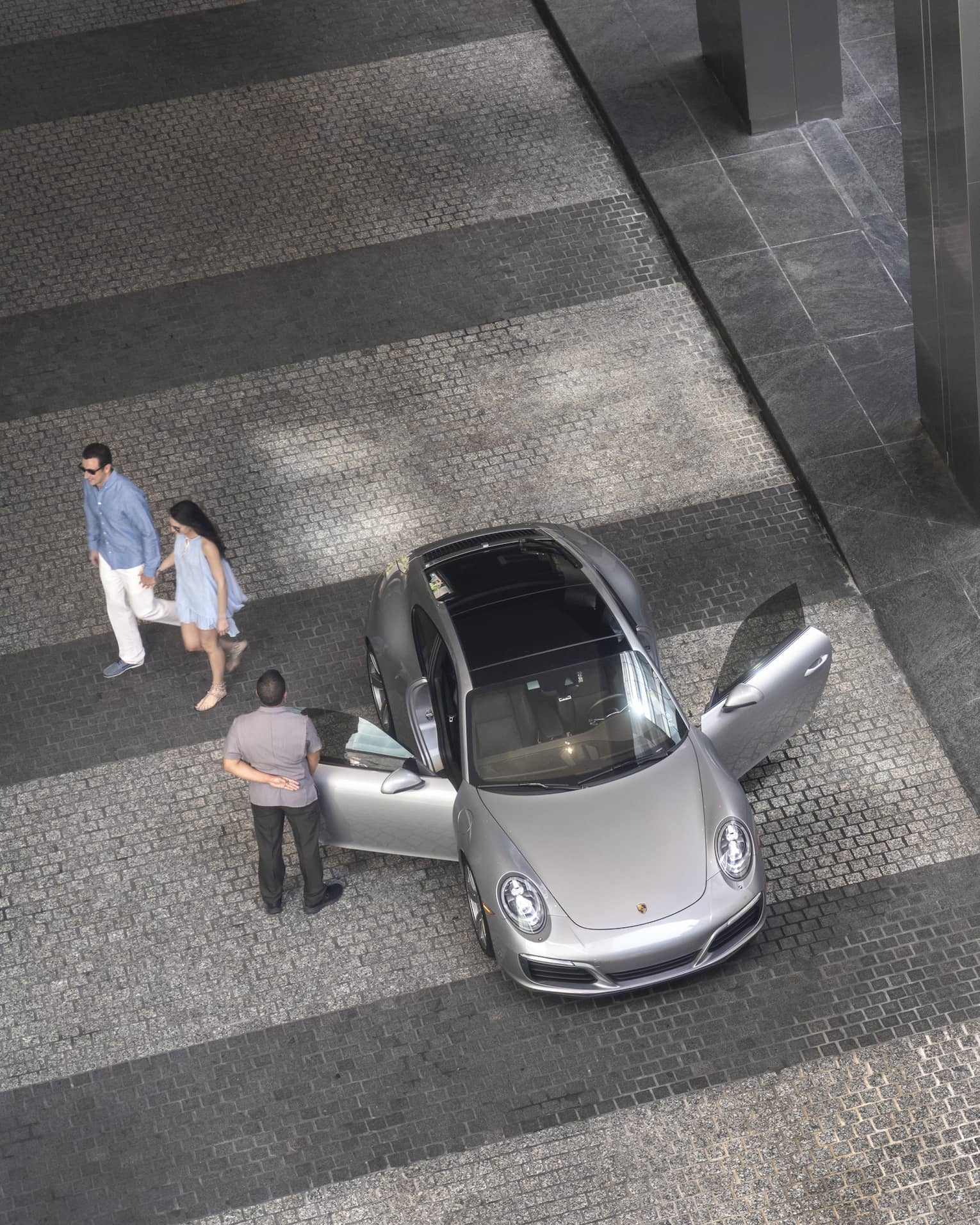 Aerial view of hotel staff opening door of luxury silver car on driveway, couple walking in background