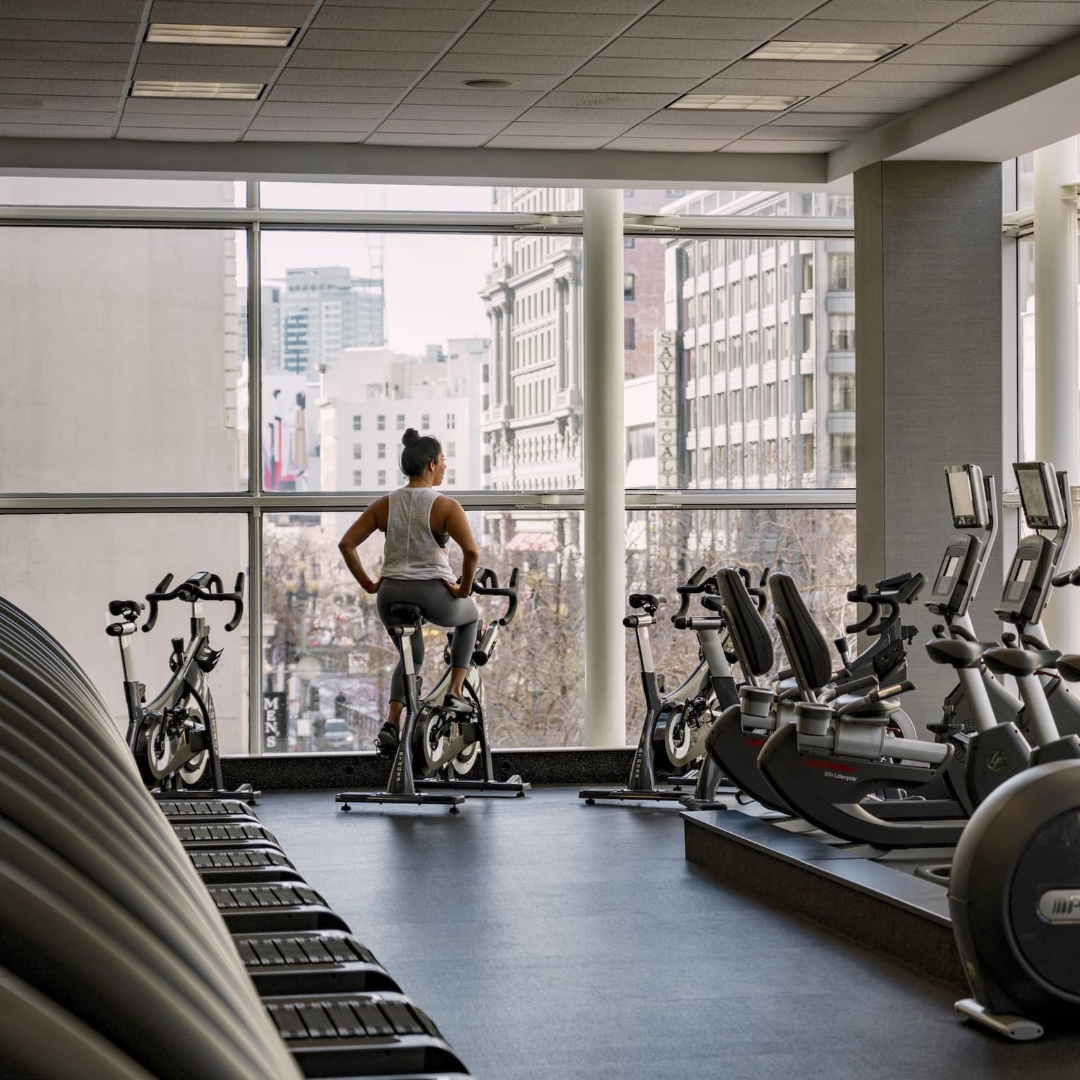 A woman rides a stationary bike in a hotel fitness center with floor-to-ceiling windows overlooking a city view