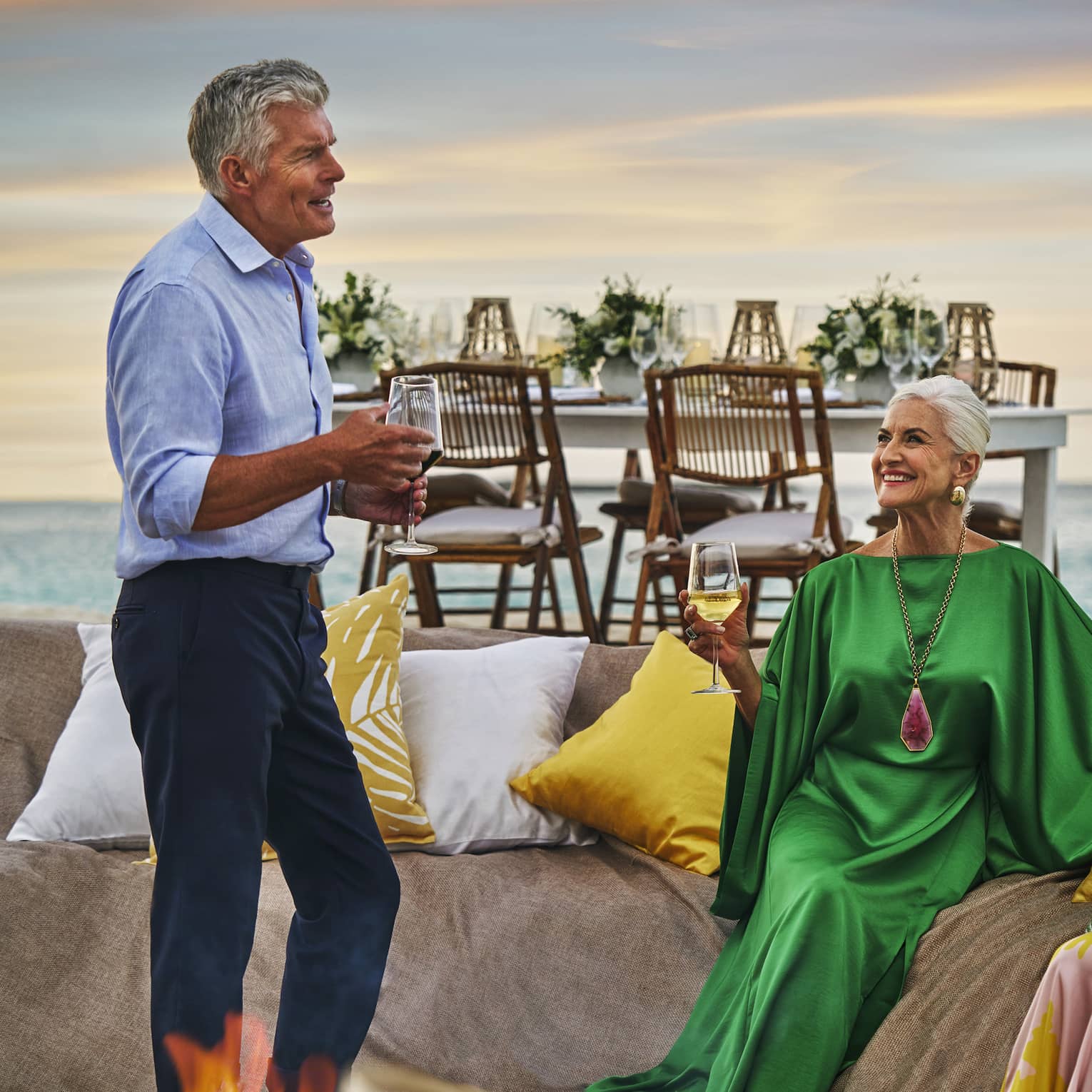 Four guests gathered around a bonfire on the beach and talking to one another.