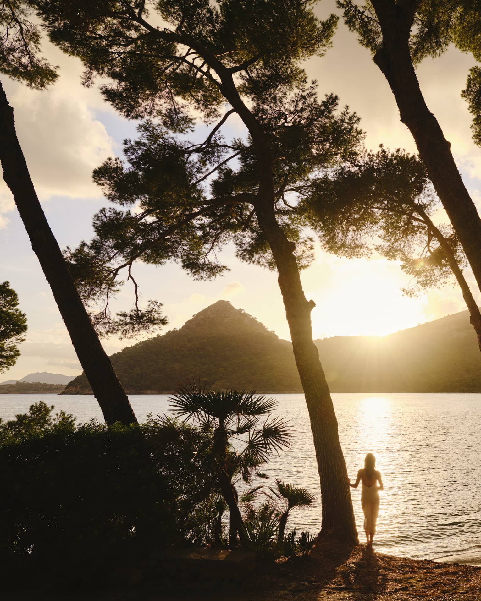 Silhouetted by the sun dipping behind a mountain, a guest stands at the edge of rippling water framed by tall leaning trees.