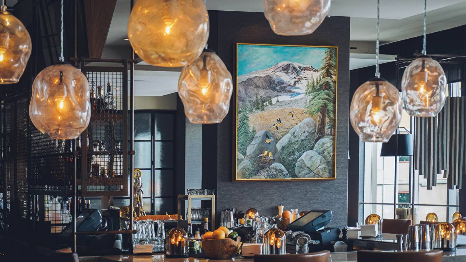 Amber-tinged glass lamps hang over bar in front of modern landscape painting in Goldfinch Tavern