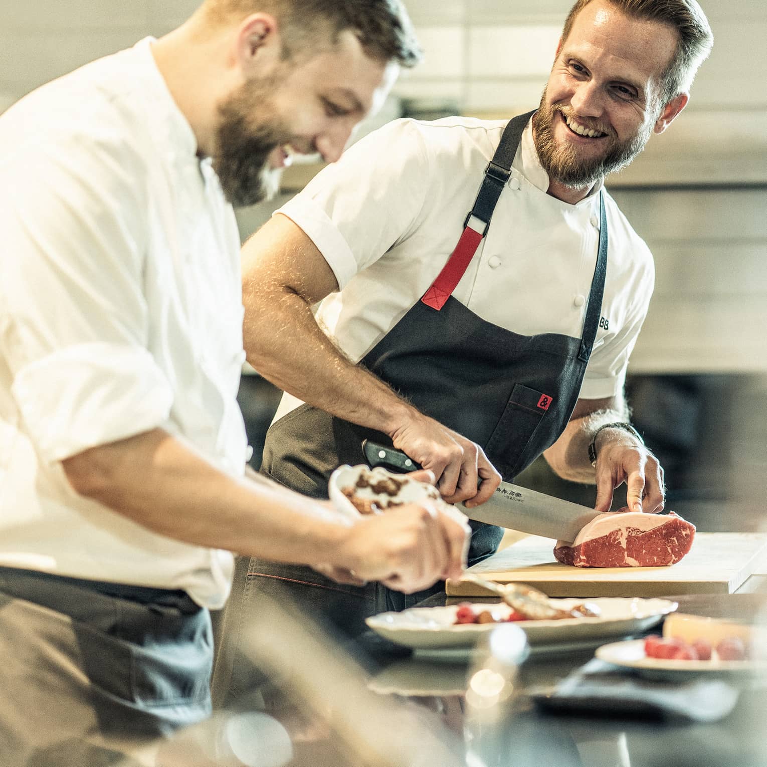 Two smiling chefs stand at a metal counter, one chopping a piece of meat while the other arranges food on a platter.