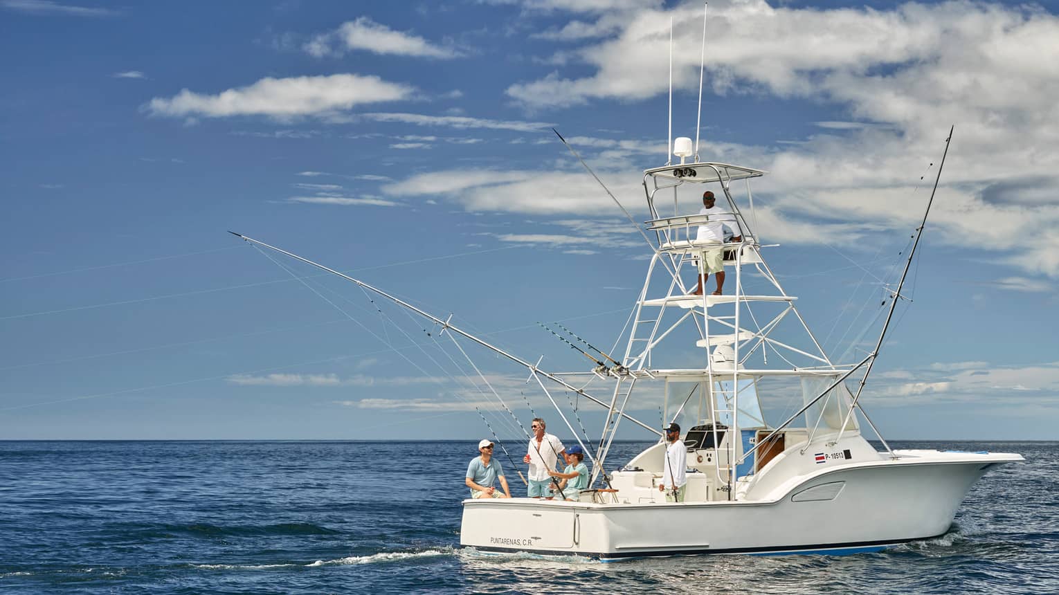 A group of people on a white fishing boat in calm ocean waters, with fishing rods ready. One person is standing on the upper deck, and the sky is partly cloudy.