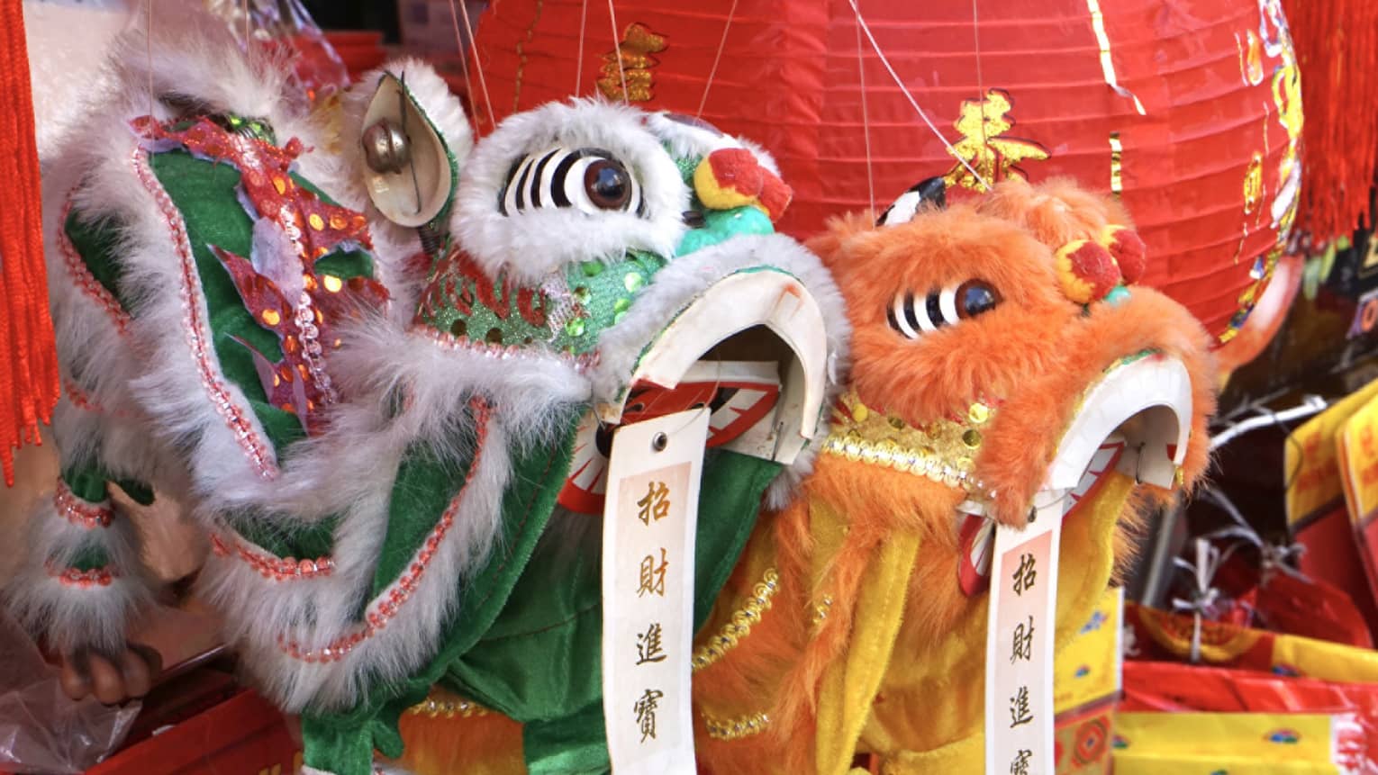 Close-up of colorful lion dance costumes in green and orange with intricate details, surrounded by red Chinese lanterns and festive decorations.