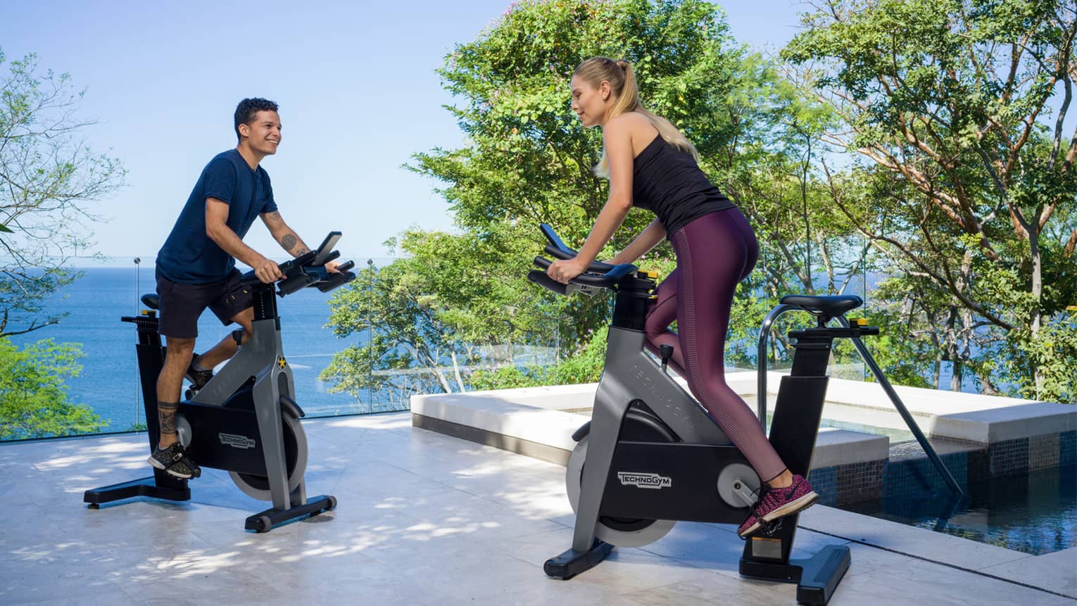 A man and woman pedal on exercise bikes on an outdoor terrace