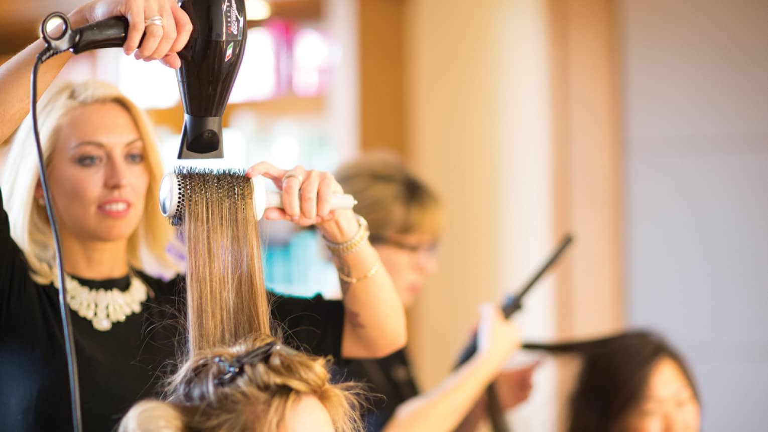 Hairstylist with hairdryer pulls back woman's hair with round brush in salon