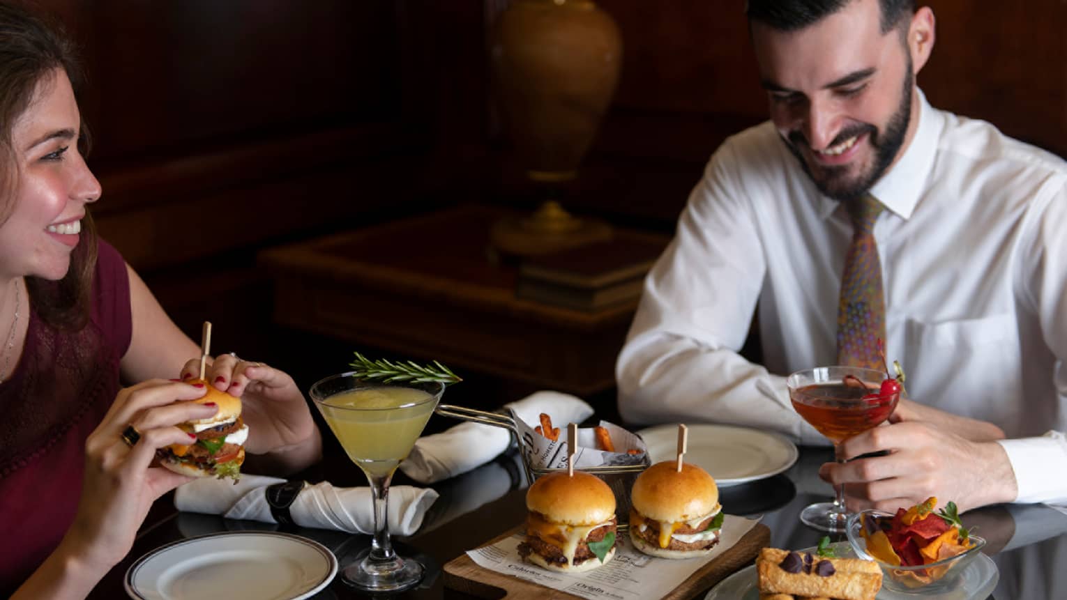 ,A couple dressed in semi-formal attire enjoys a spread of hamburger sliders, deep fried appetizers and cocktails
