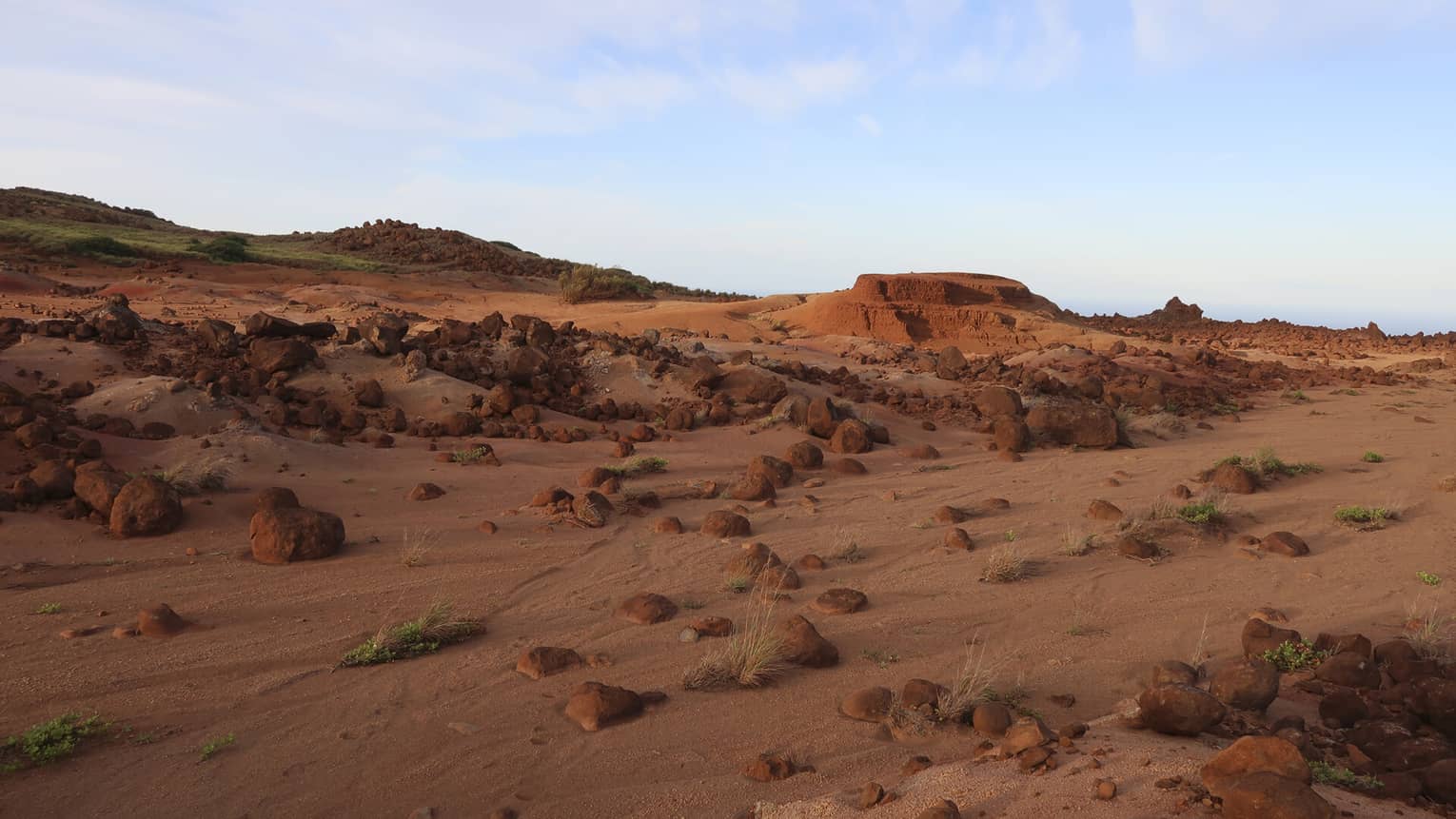 A desert landscape of red boulders, rocks, sand and small rocky hills with the occasional green vegetation growing.