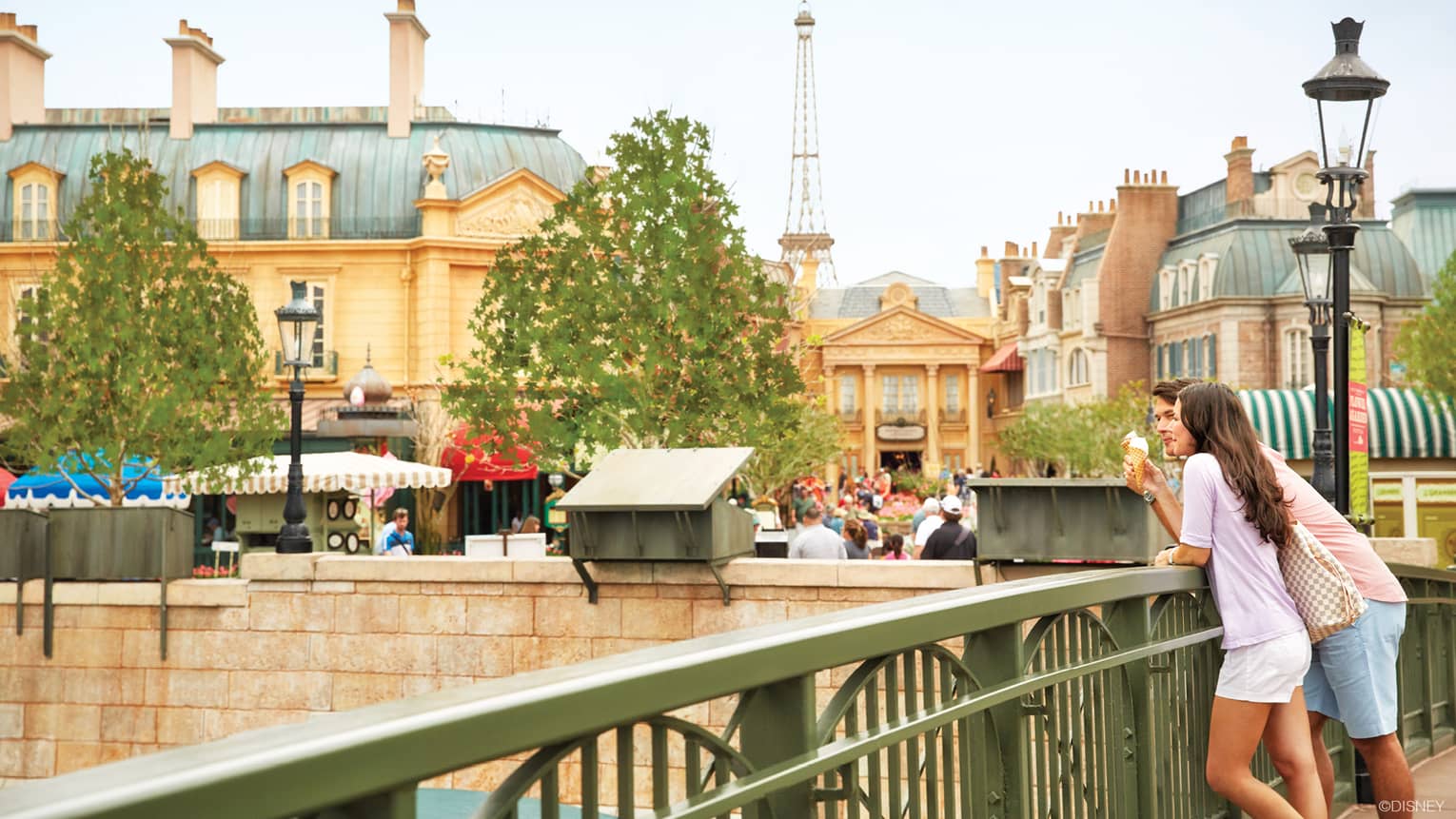 A man and woman leaning on a bridge railing while eating icecream.