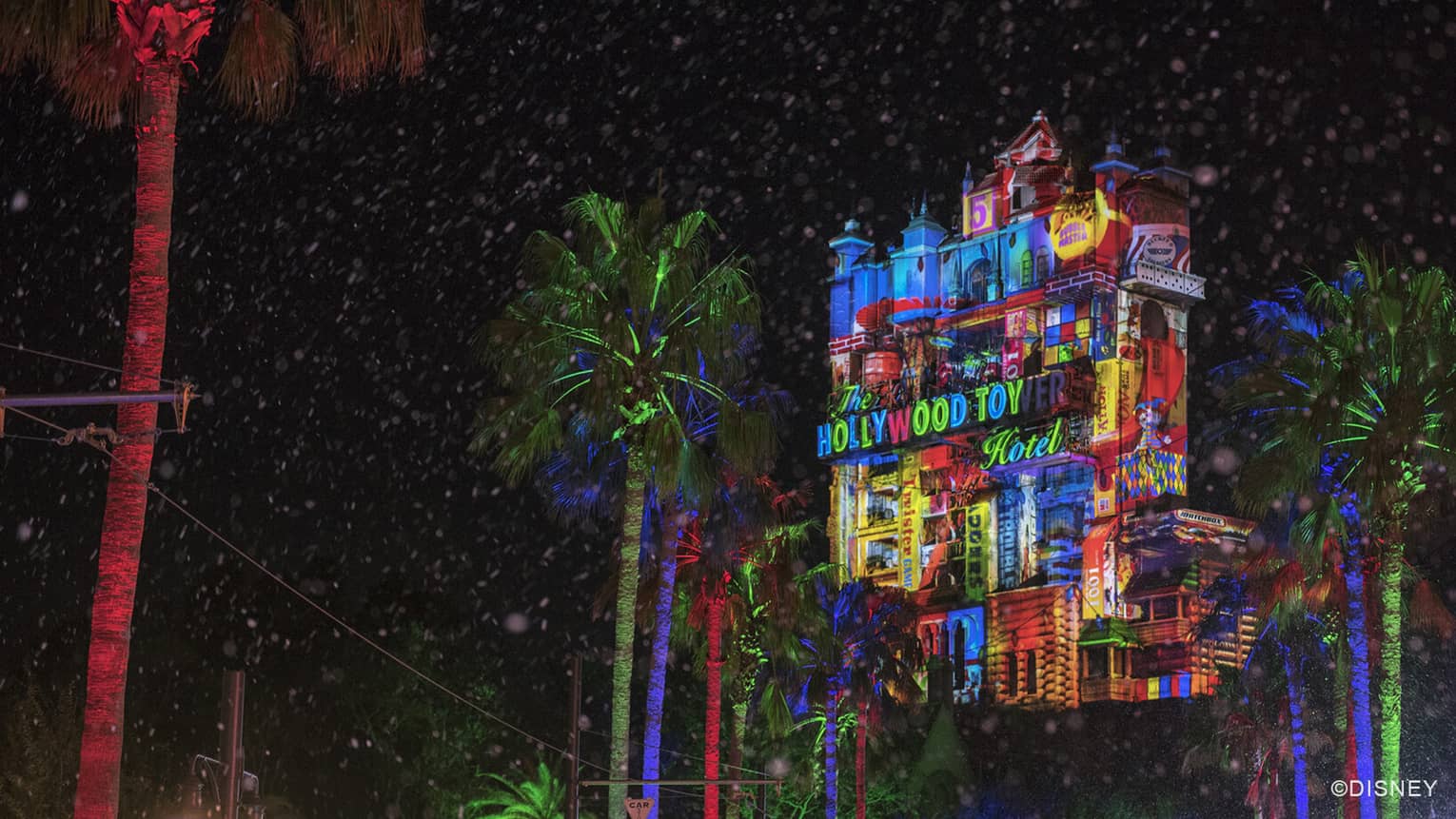 A view of Disney's Hollywood Tower Hotel ride lit up at night, with colorful festive lights and red and green lit trees below 