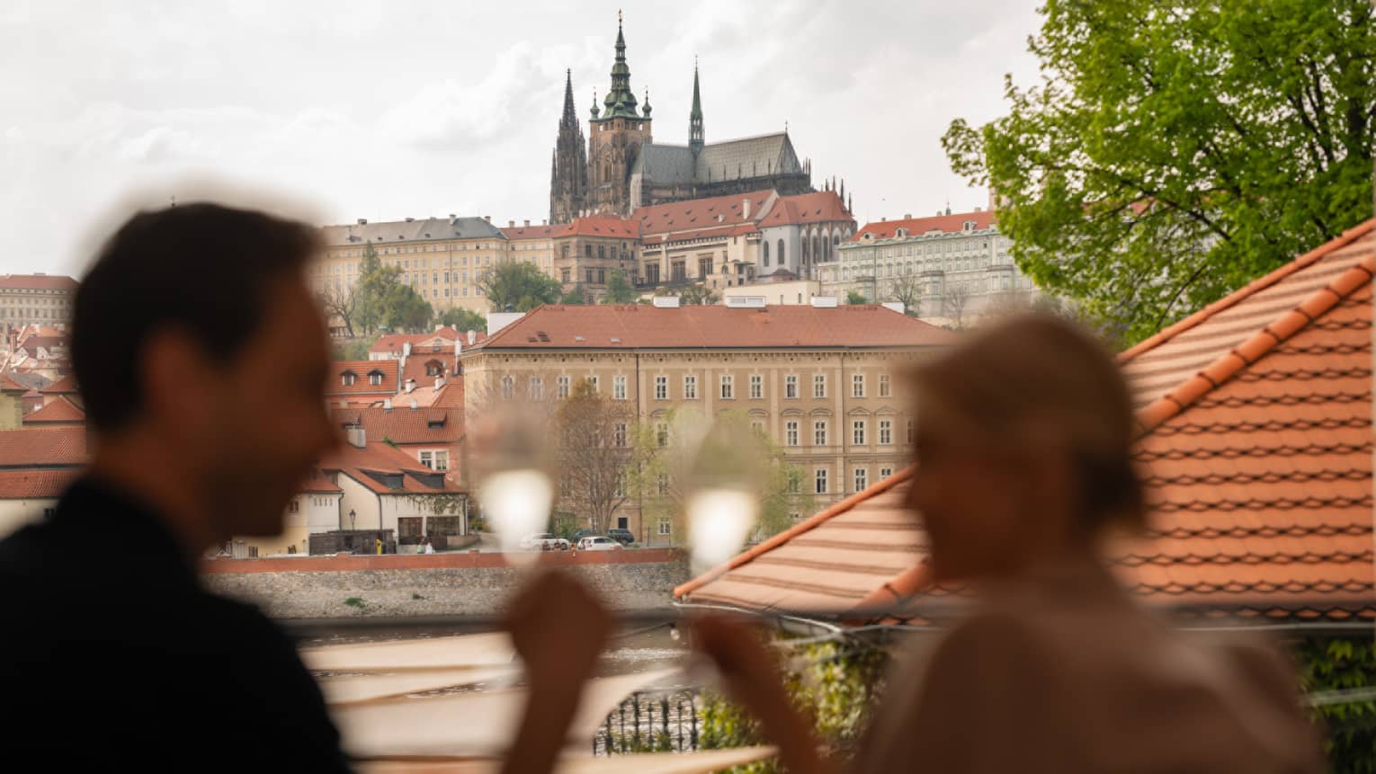 Two shadowed people on terrace, toasting with champagne over view of Prague Castle