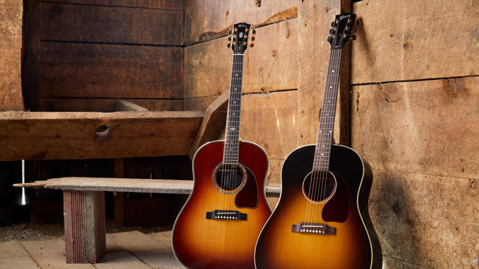 Two beautiful Gibson guitars placed on stands and set in front of an old worn-down wooden bench in a dusty rustic room.