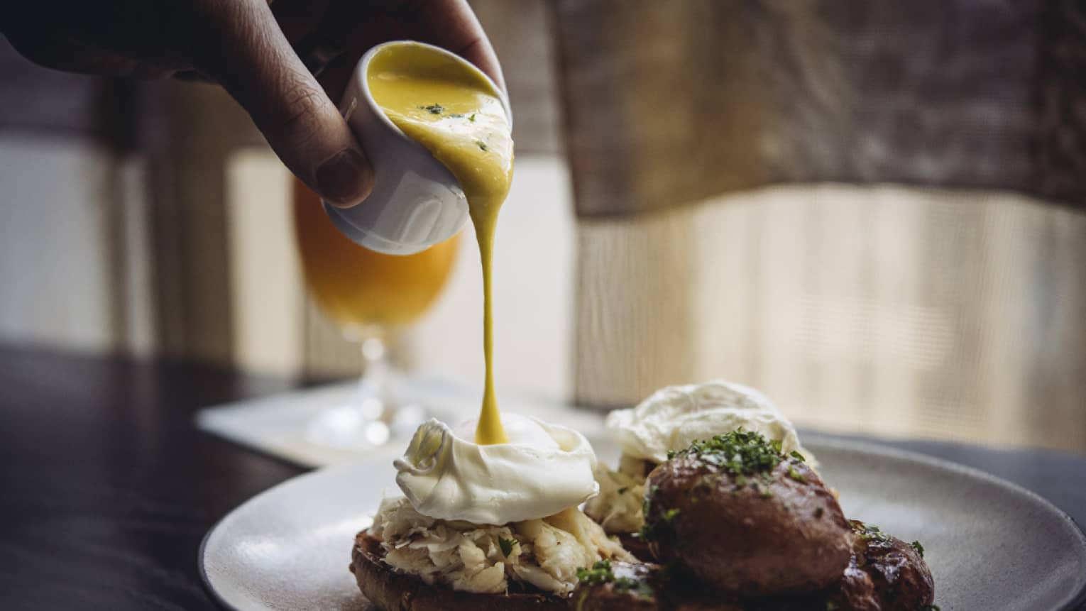 Hollandaise sauce being poured onto eggs Benedict with lump crab meat, side of crispy roasted potatoes 