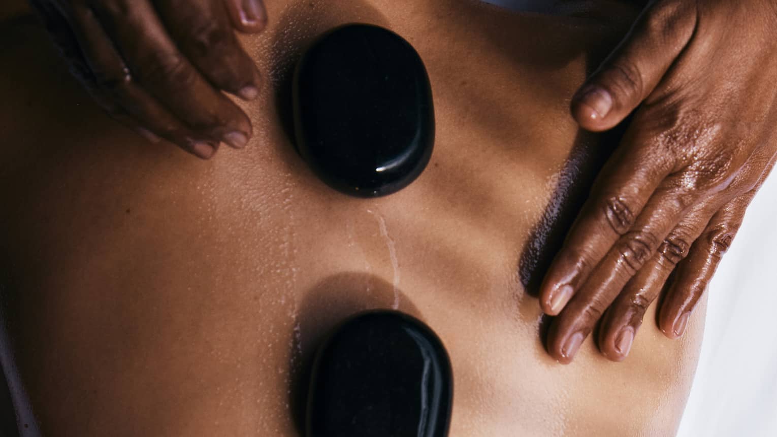 A person lying prone on a spa treatment table with smooth, round, black oval stones placed along their spine
