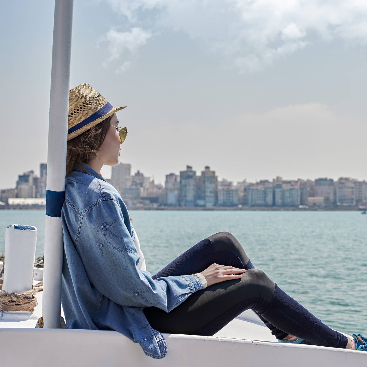 A guest wearing a straw hat sits on the bow of a boat and looks out over calm water toward a distant metropolitan shoreline.