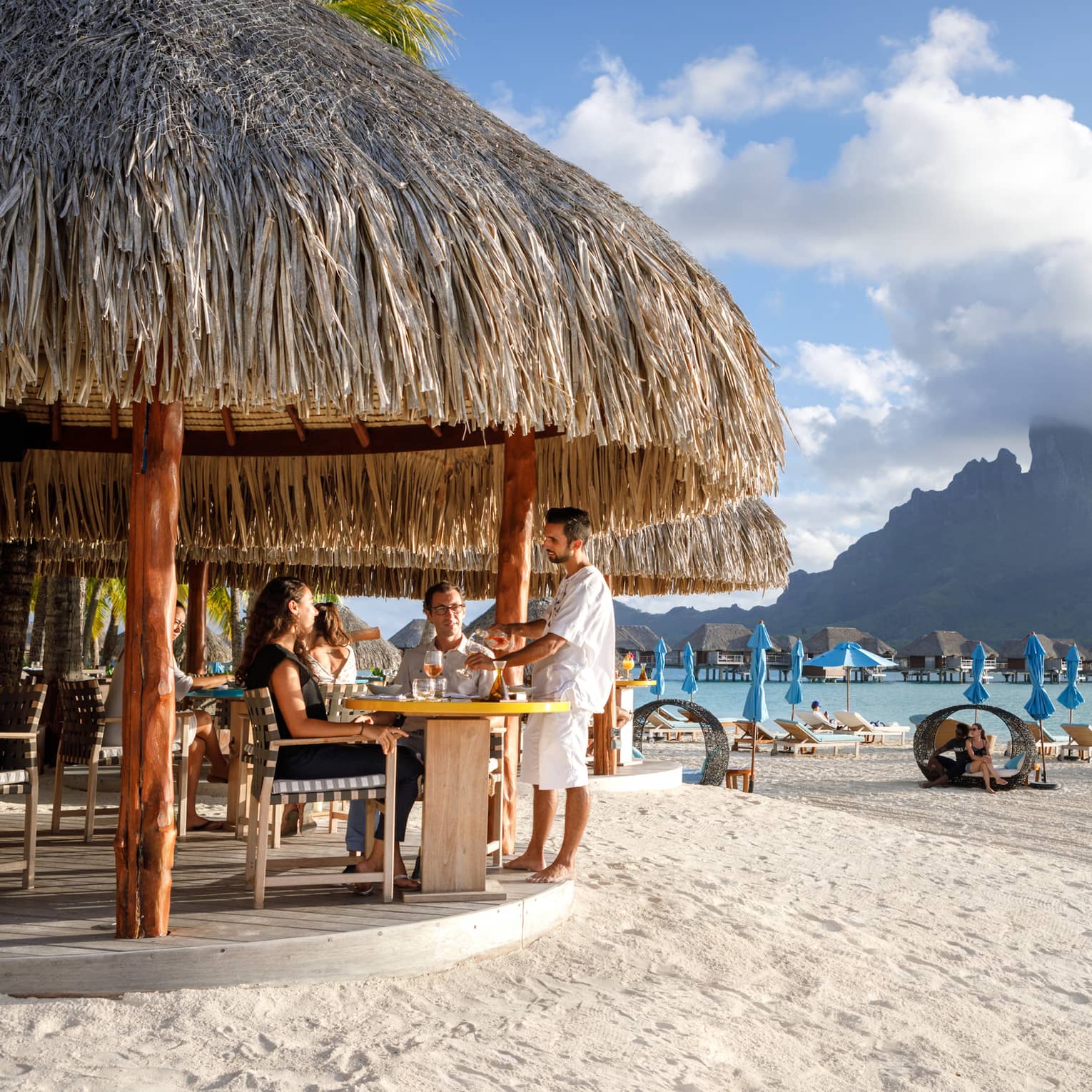 Couple seated in bar under grass roof on beach, view of people in personal cabanas, mountain view