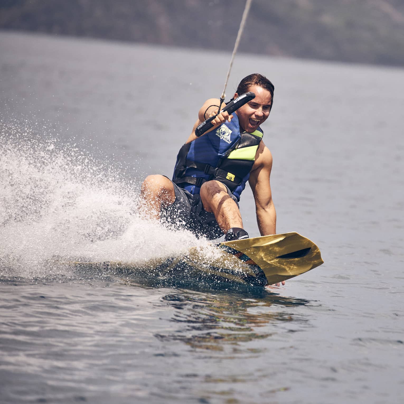 Young man wearing a blue and green life vest and blue swim trunks wakeboarding through the water