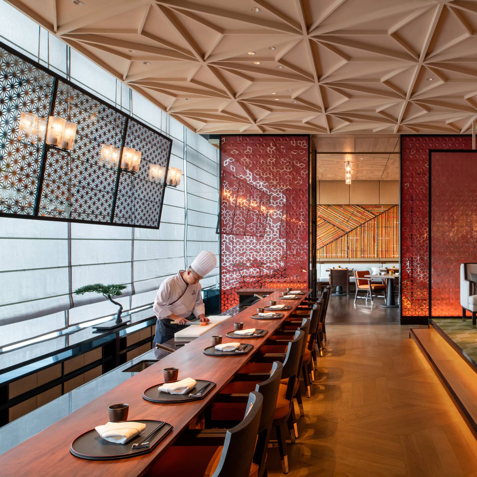 A sushi chef works by a long wood dining table in a modern restaurant, a red ornate screen and walls reflecting golden light.