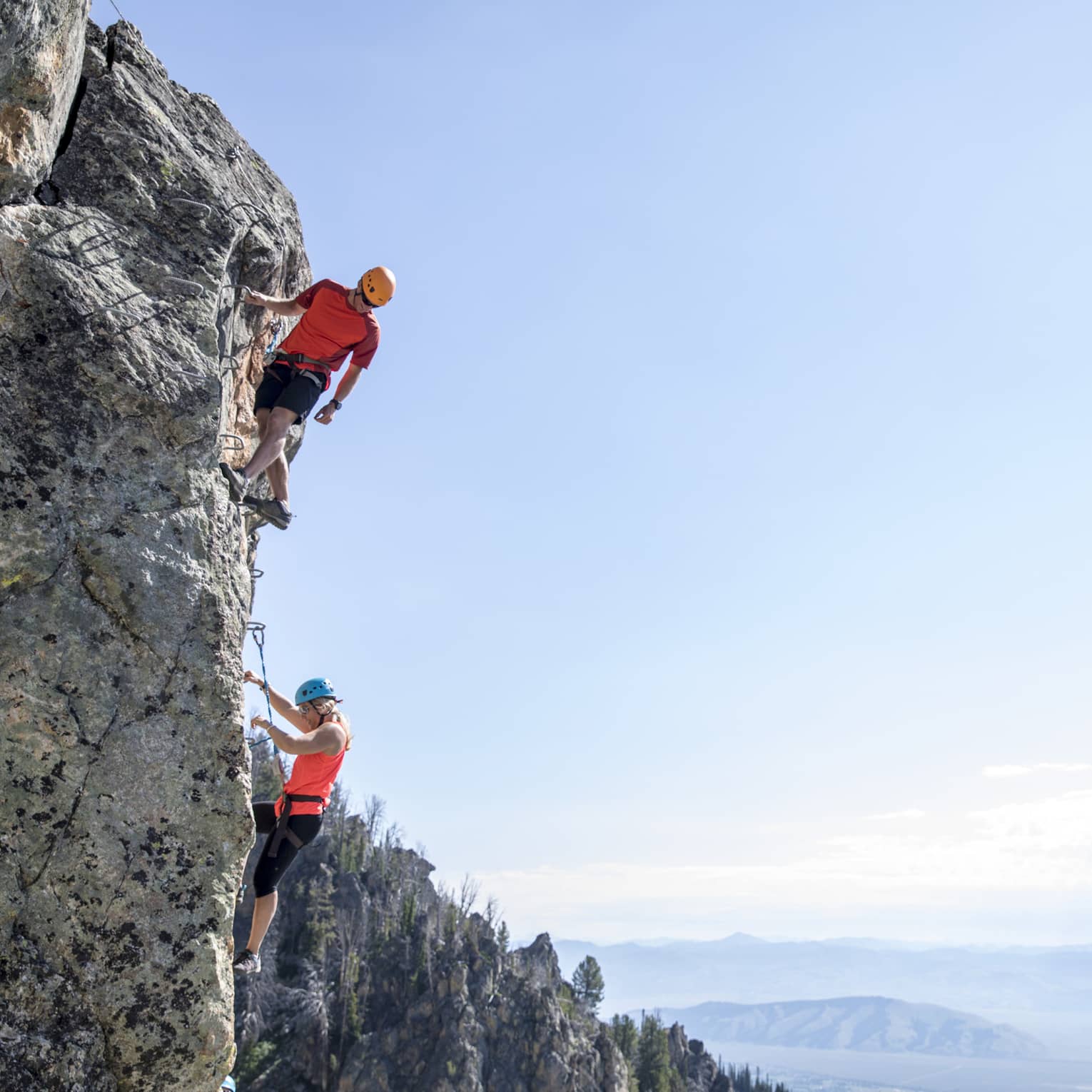 Man and woman climb steep cliff face with view of mountains in far distance 
