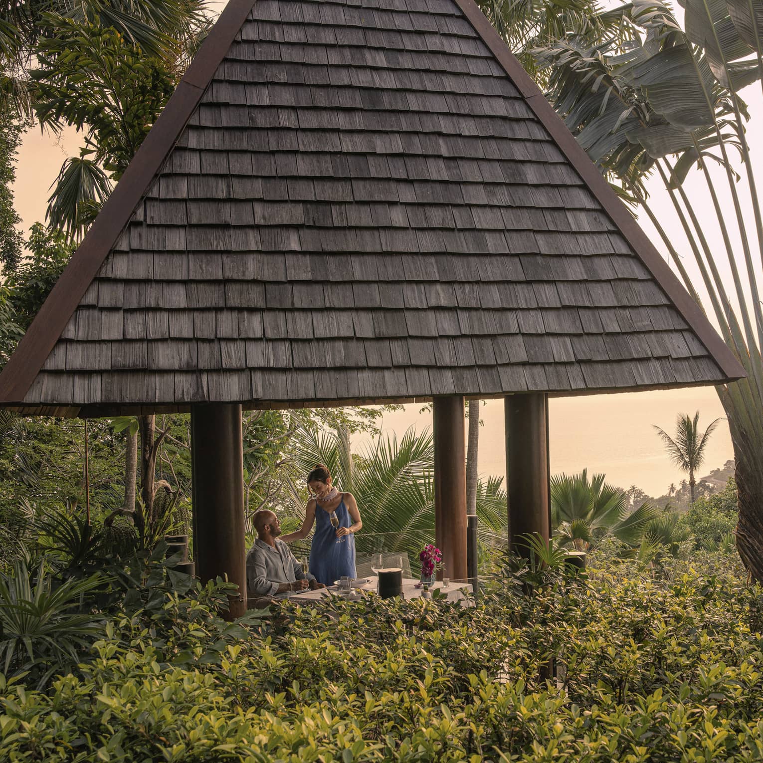 A couple with champagne in a tall hilltop pavilion with a dark, straw roof, set amidst lush forest and a giant fan palm tree.