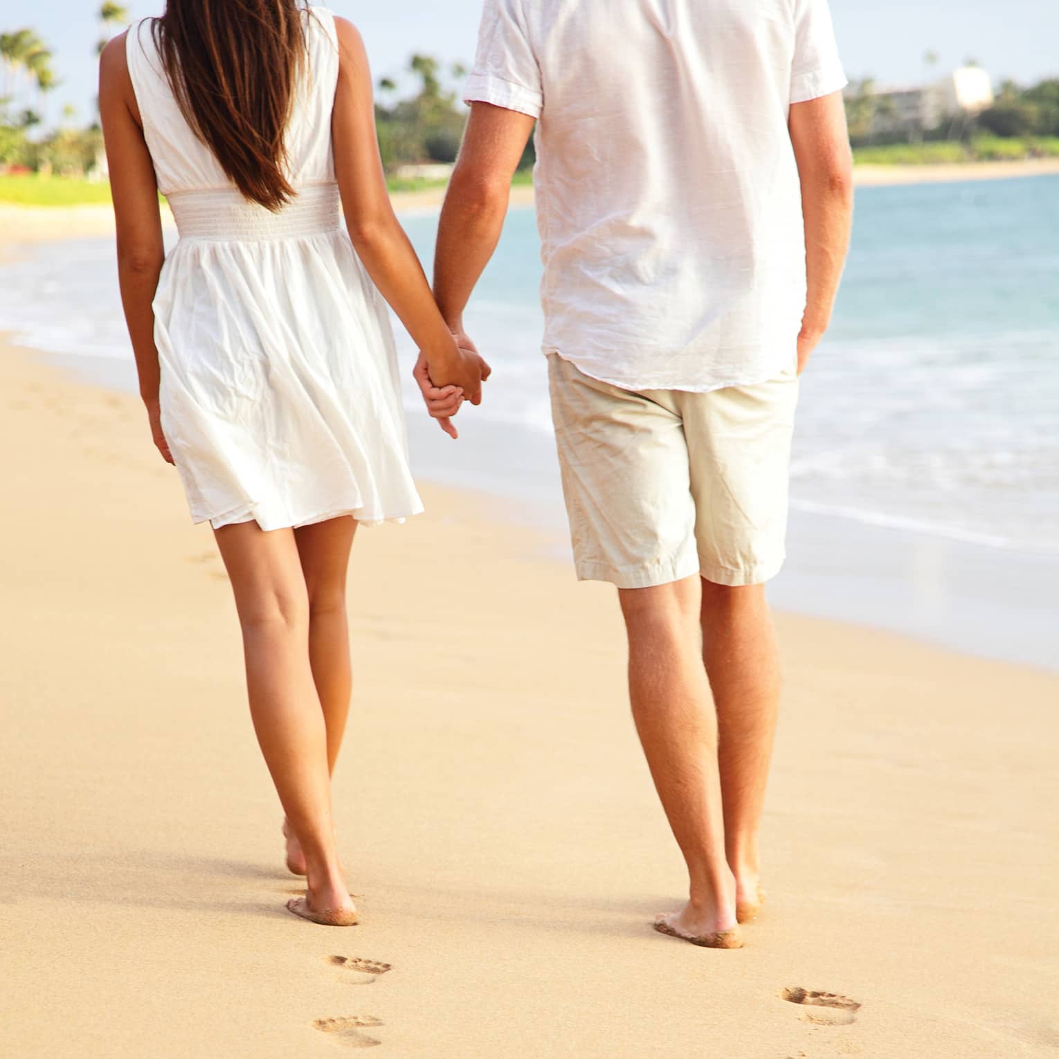 A close-up of a couple holding hands and leaving a trail of footprints as they stroll down a beach near the water’s edge.