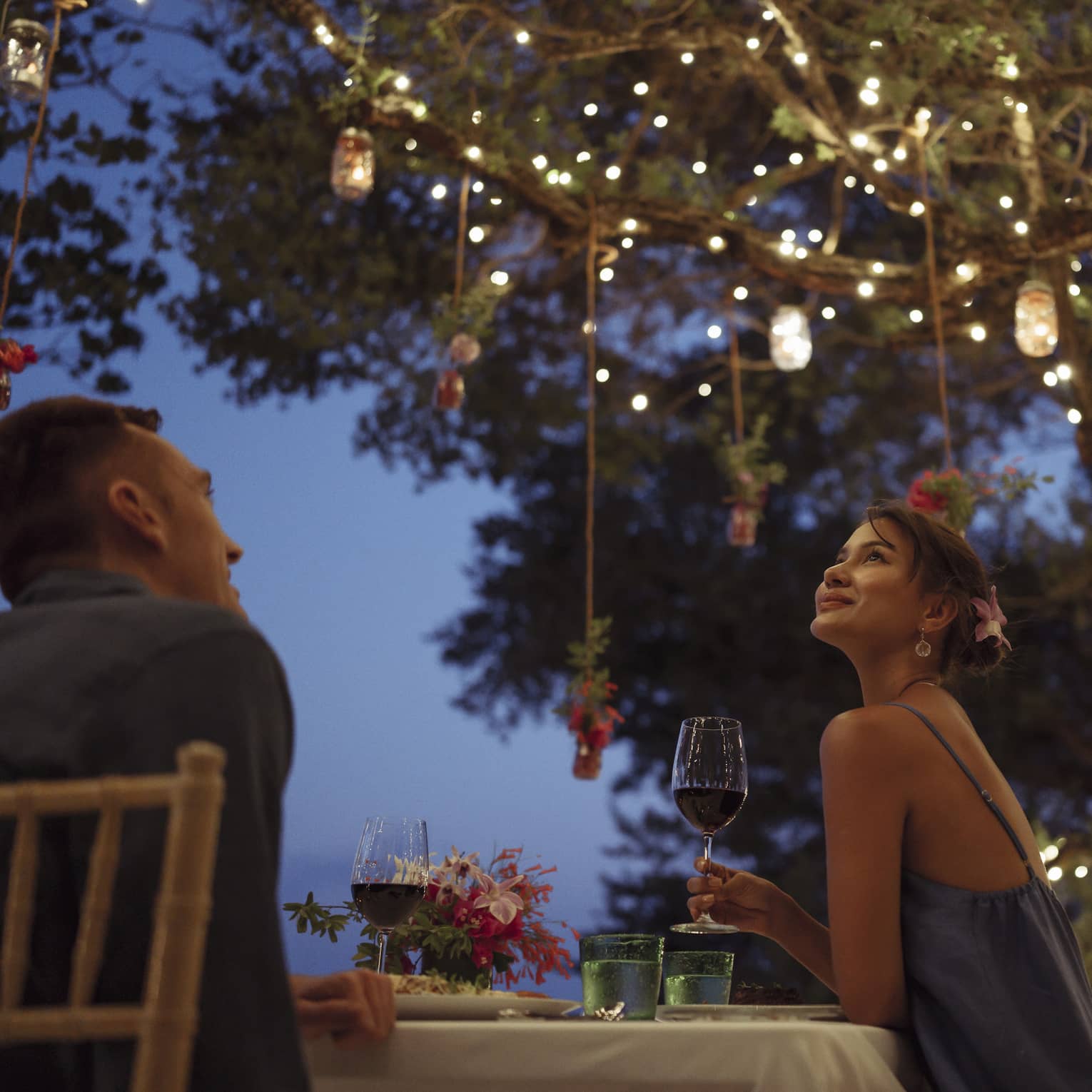 A smiling couple having a romantic dinner under trees covered in string lights and jars of flowers hanging from the branches.