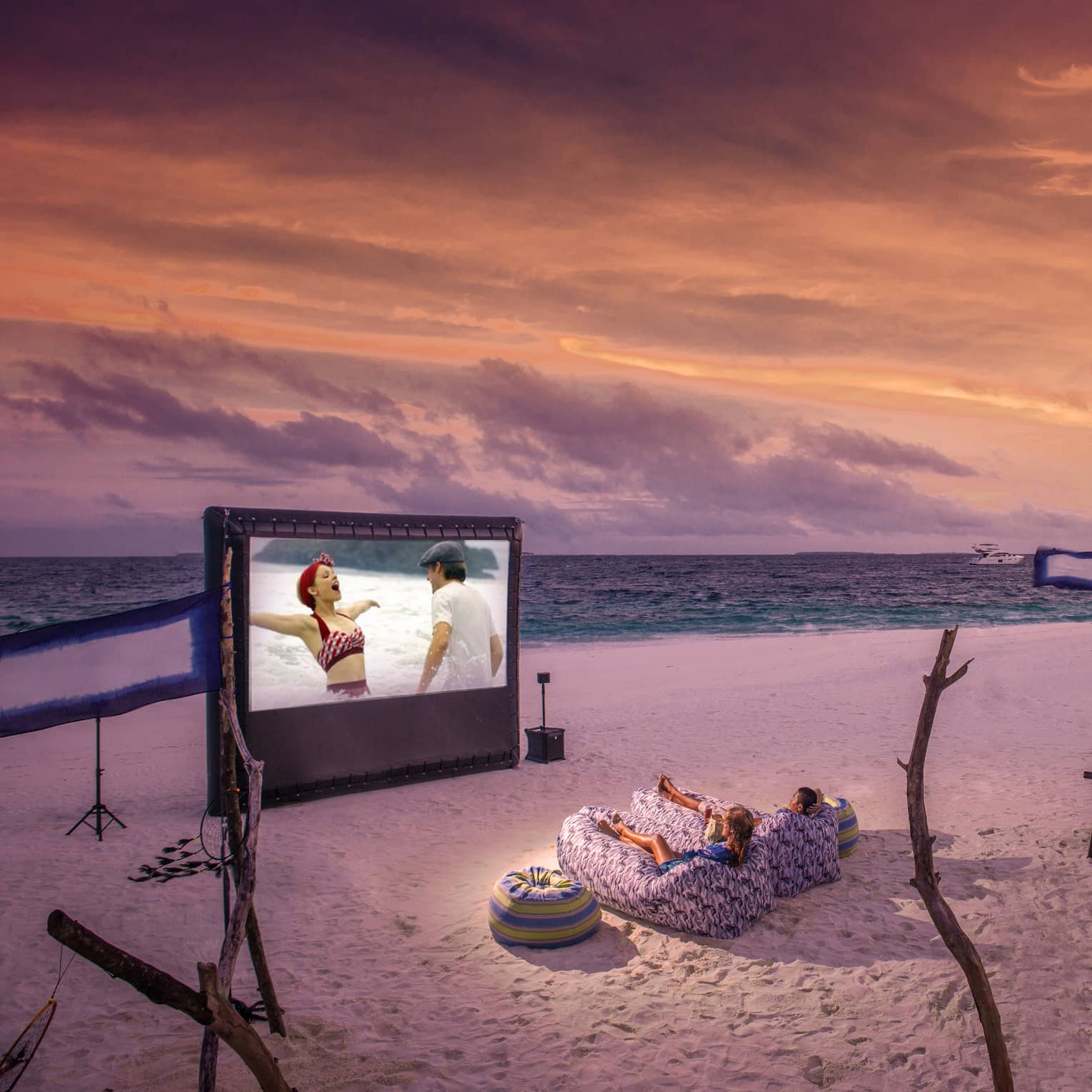 ,Two people in beanbag chairs enjoy a movie night on the beach at dusk, popcorn stand on sand behind them