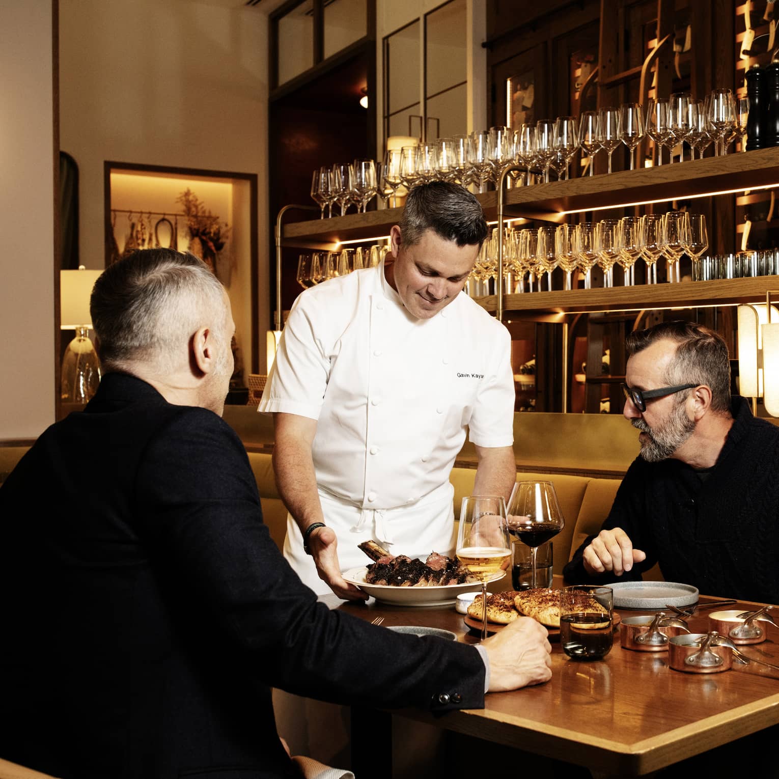 Chef brings a plate of food to two people seated at a table in front of a wood-and-brass shelf of wine glasses