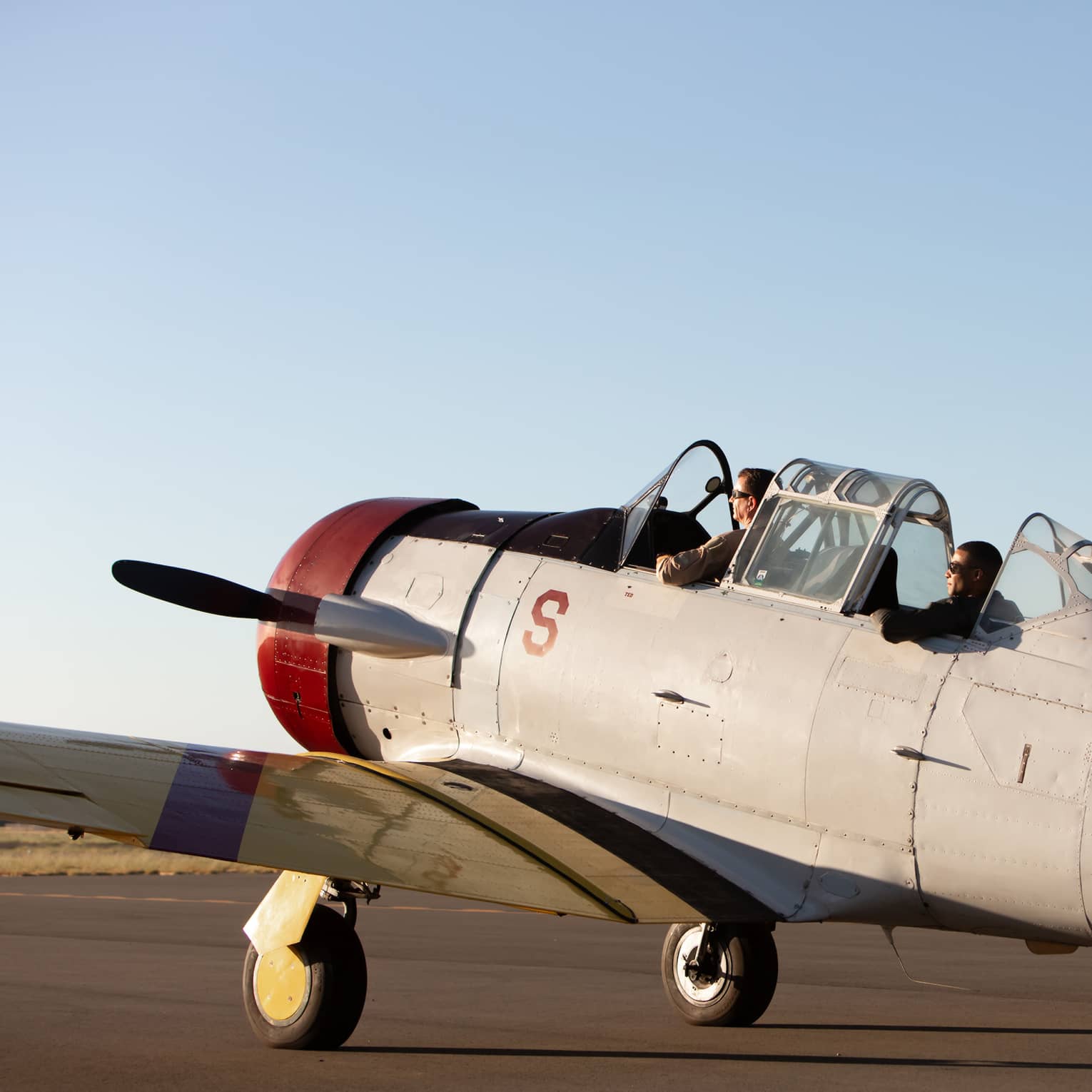 Two people sit in the cockpit of a vintage U.S. Navy aircraft on a runway, preparing for takeoff under a clear sky.