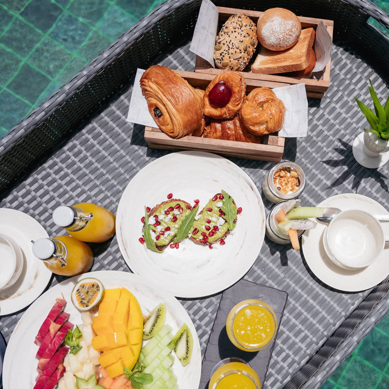 Close-up view from above of a wicker tray filled with fruit, pastries, juice and toast floating in a crystal-clear pool.